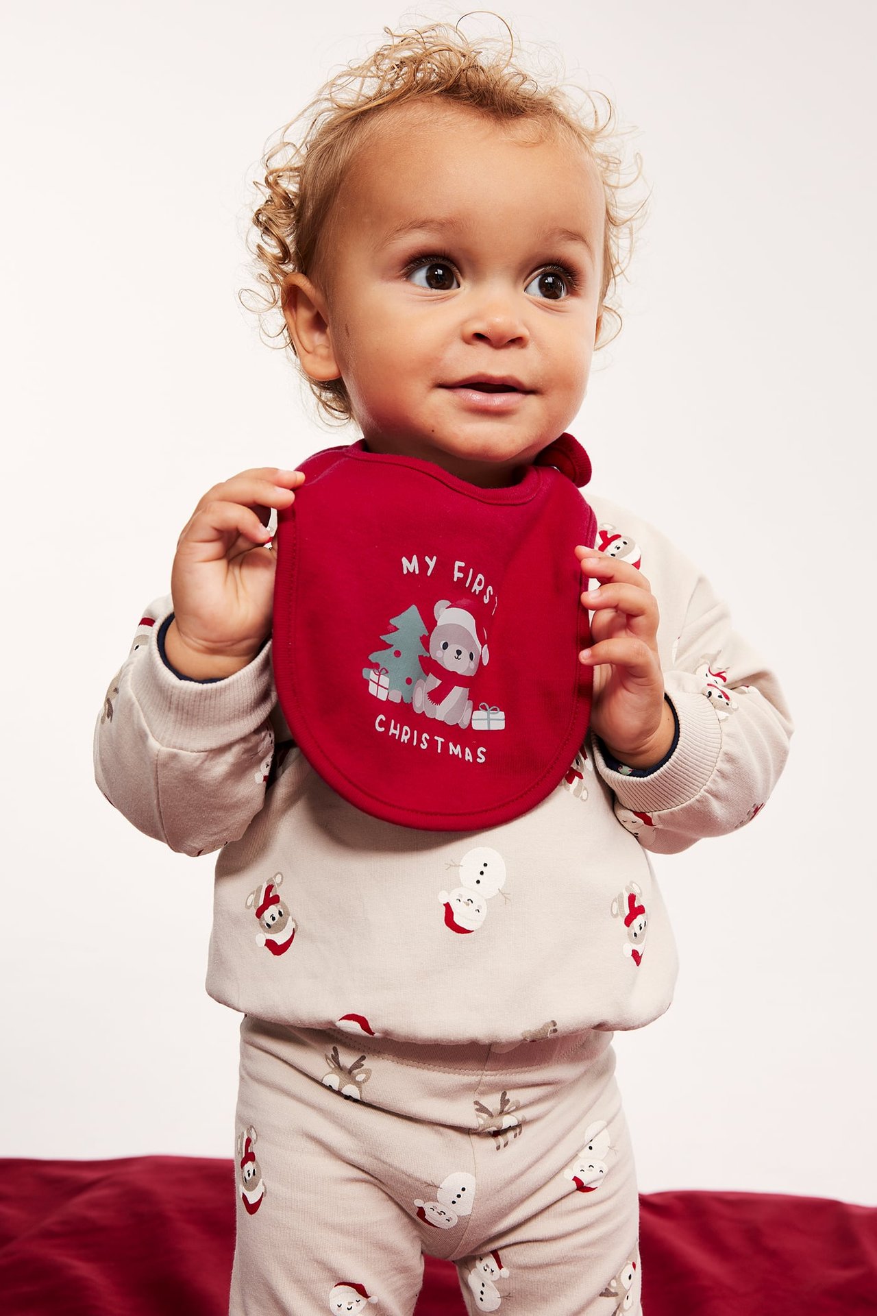 Baby trzyma czerwony śliniak 'My First Christmas', ma na sobie beżową piżamę w świąteczny wzór.