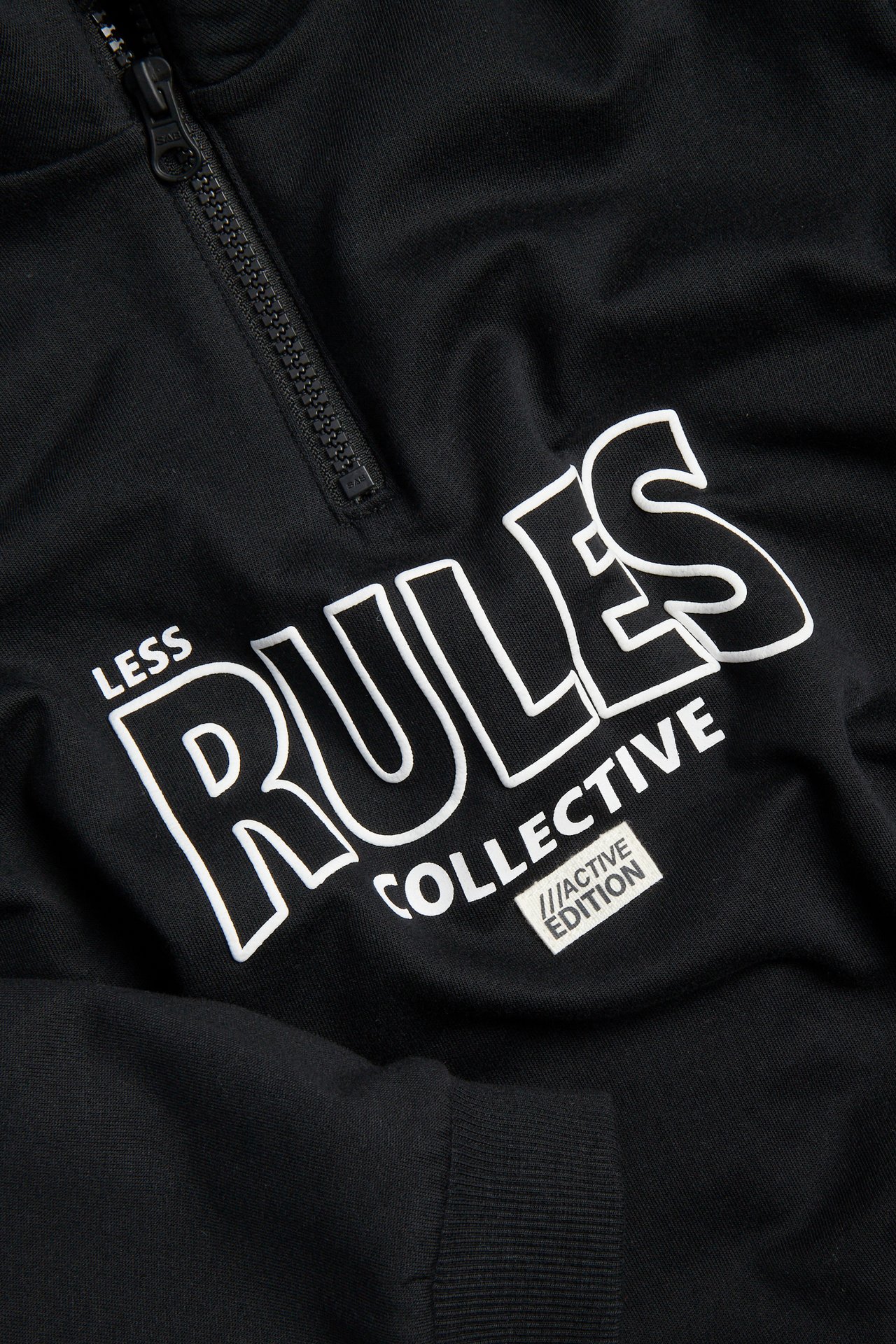 Närbild på en svart collegetröja med kort dragkedja och vitt 'LESS RULES COLLECTIVE'-tryck på bröstet.