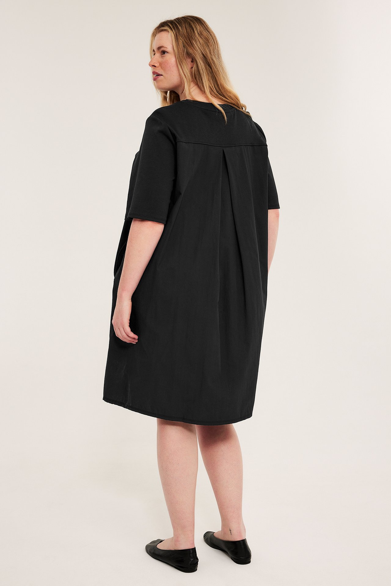 Bakifrån-sida vy: kvinna i svart, ledig t-shirtklänning med veck i ryggen och svarta ballerinaskor.