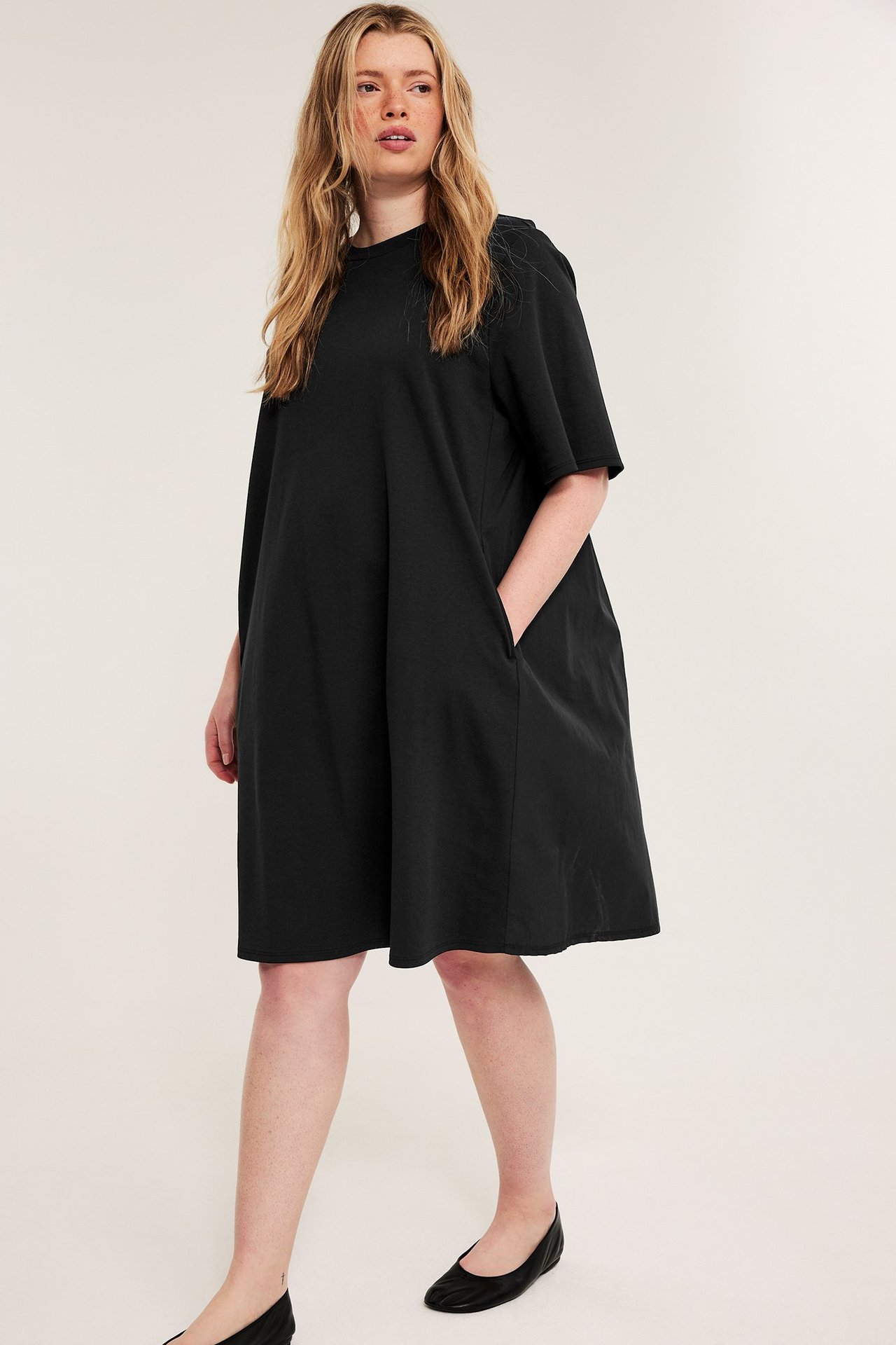 Framsida: kvinna i svart avslappnad kort t-shirtklänning med fickor och svarta ballerinaskor.