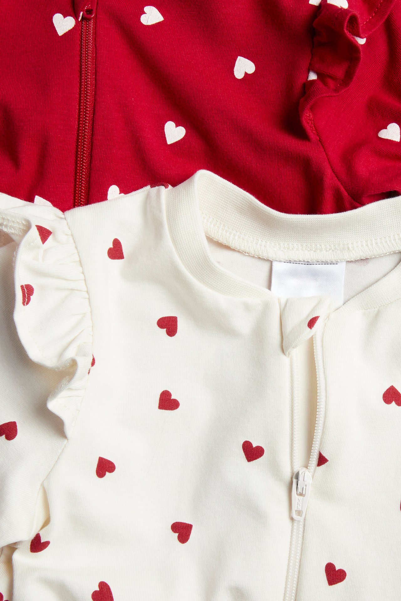 Vit babybody med röda hjärtan och volanger, dragkedja fram, bredvid rött babyplagg.