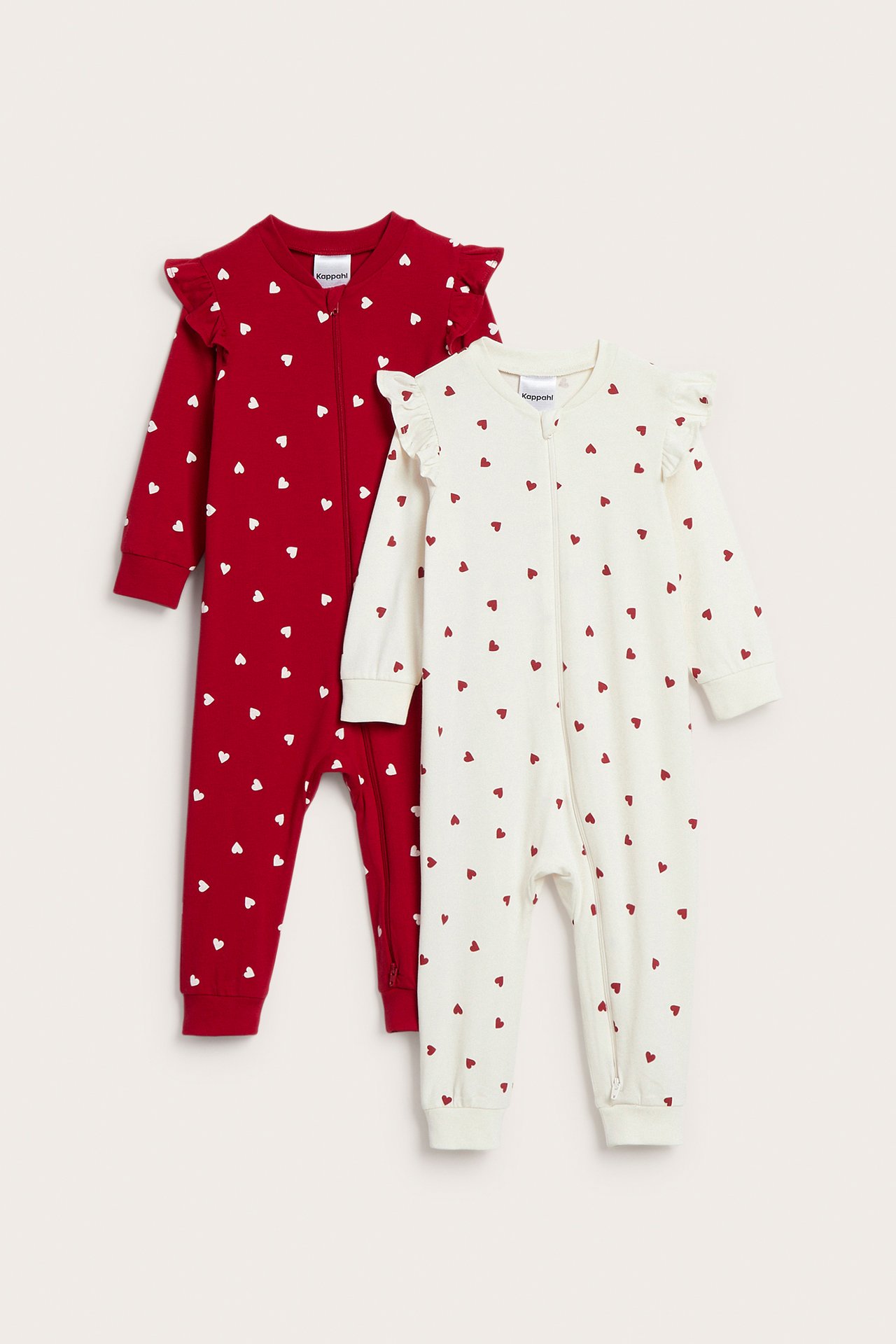 Framifrån, två baby-sparkdräkter med volanger: en röd med vita hjärtan, och en naturvit med röda hjärtan.