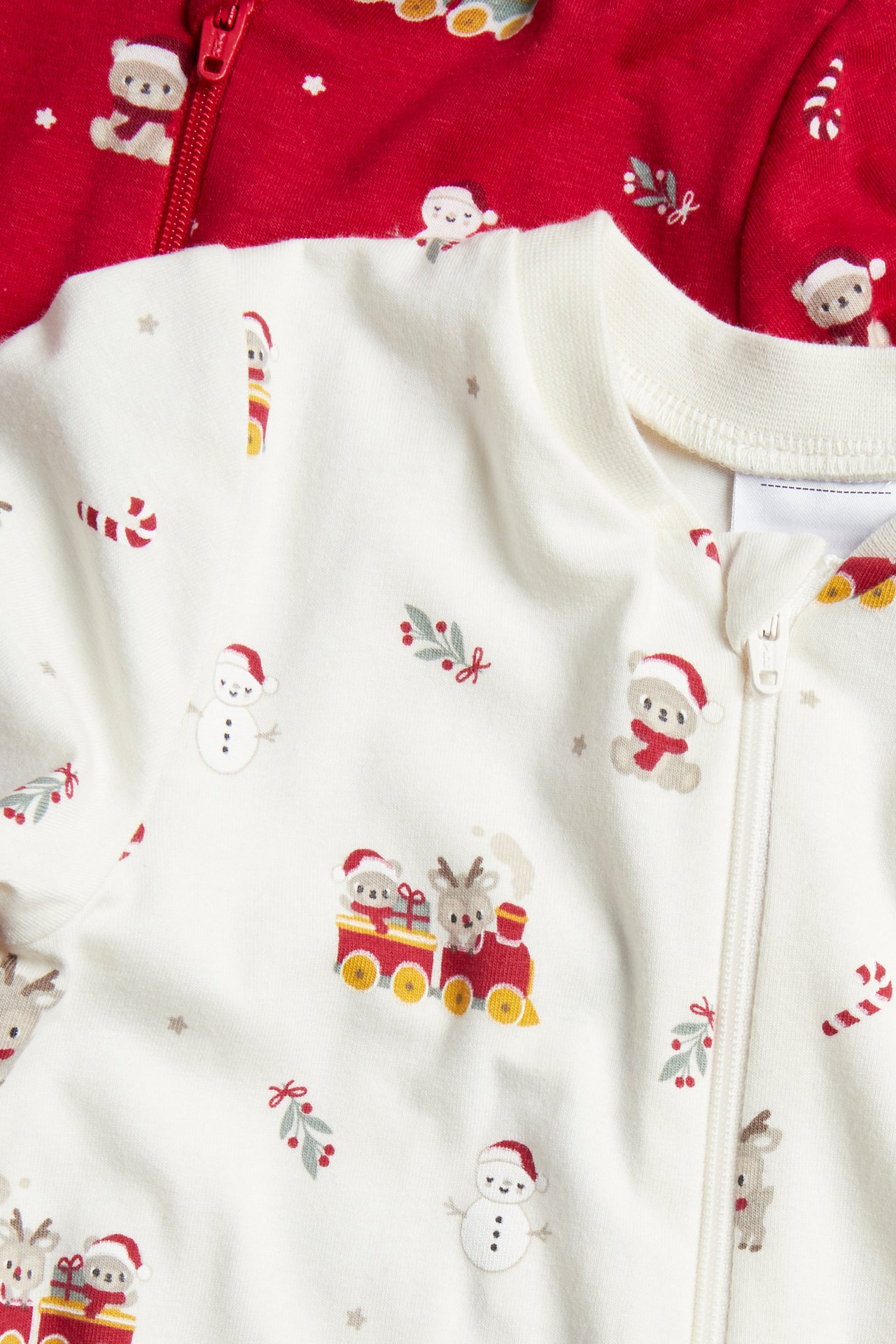 Närbild, framifrån på en krämfärgad babypyjamas med julmönster och dragkedja, bredvid en röd pyjamas.