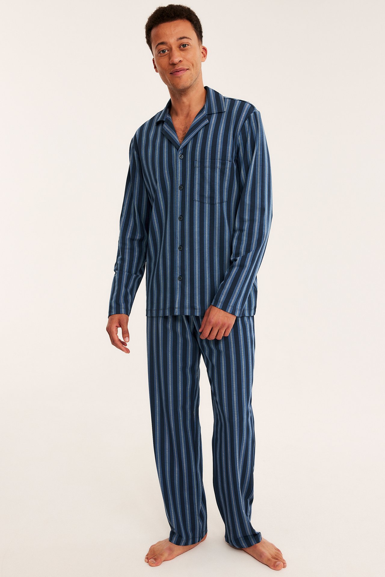 Edestä kuvattuna miesmalli yllään sinisen ja vaaleansinisen raidallinen pitkähihainen pyjamas napeilla.
