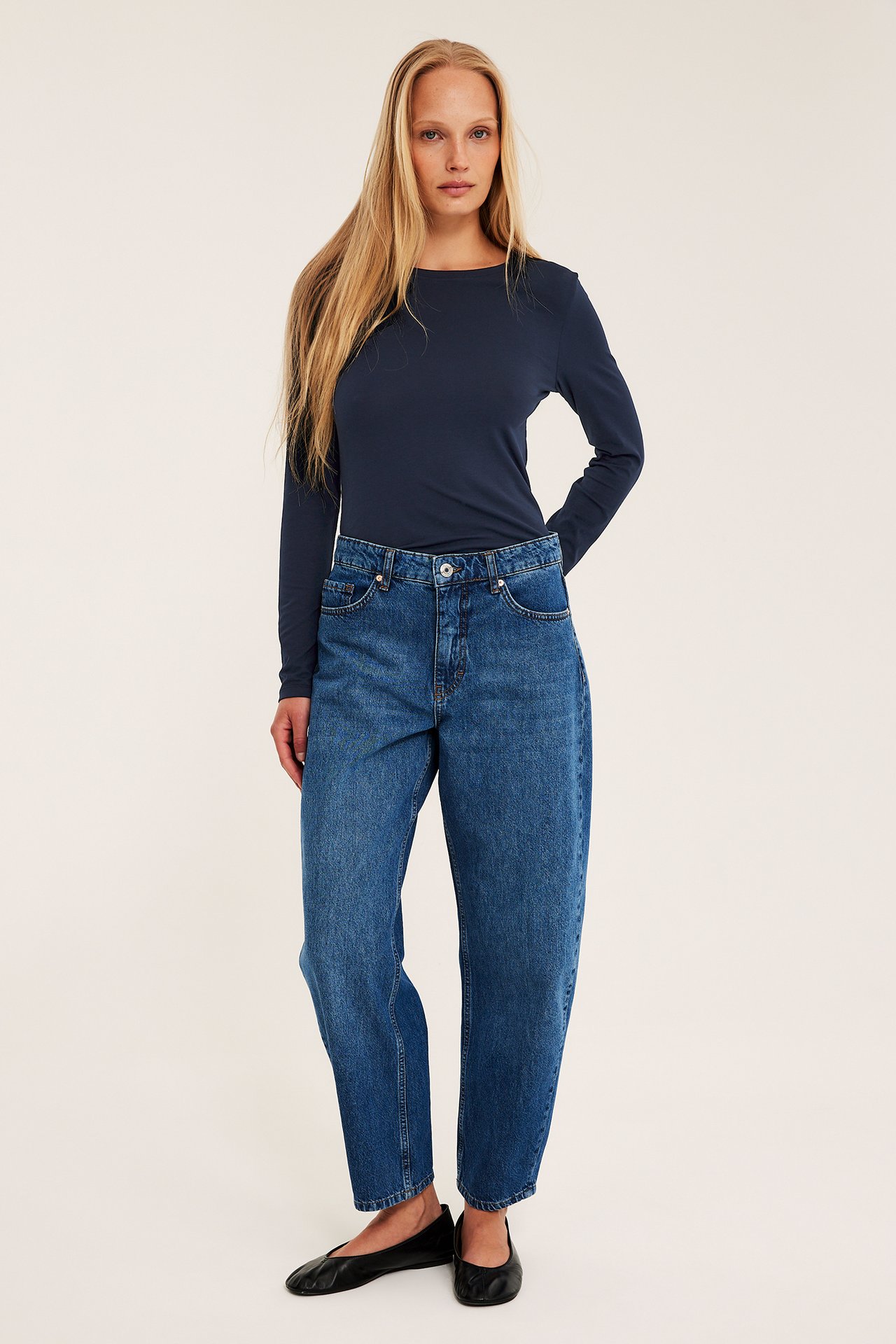 Framifrån: kvinna i mörkblå långärmad topp med rundhals, blå höga jeans och svarta platta skor.