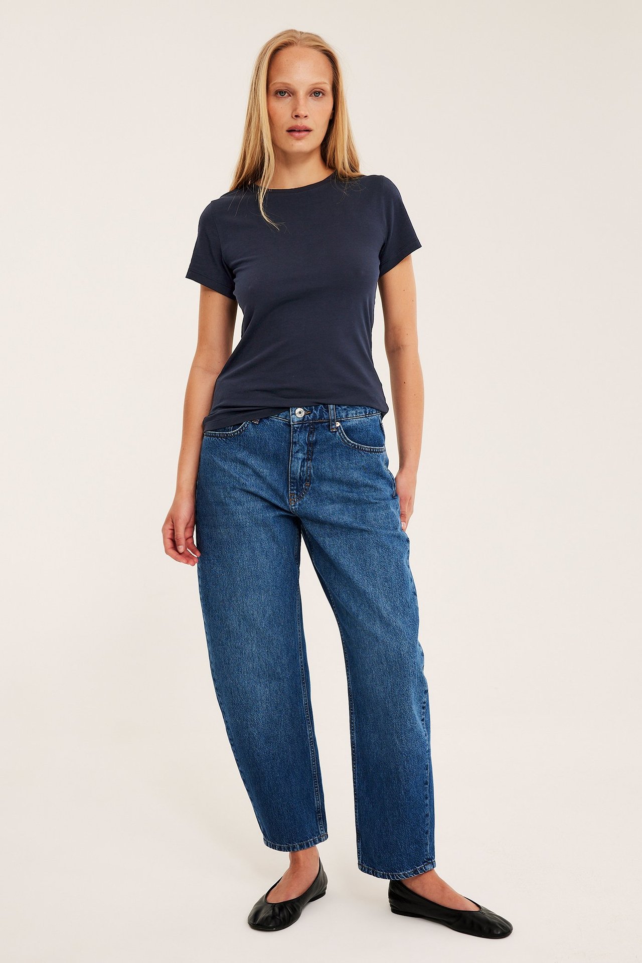 Forfra: kvinne har på seg en mørkeblå figurnær kortermet t-skjorte, blå jeans og svarte ballerinasko.