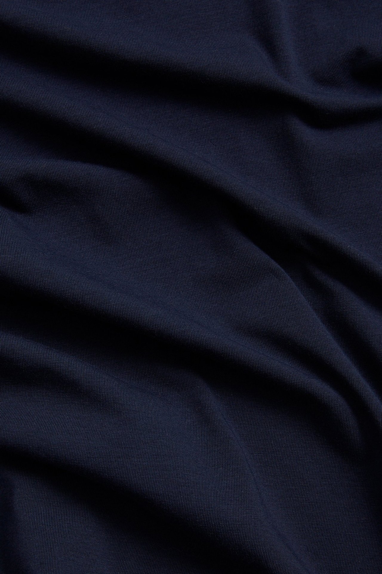 Nærbilde av mørkeblått, ensfarget bomullsblandingsstoff fra en figurnær t-skjorte, med myke folder.