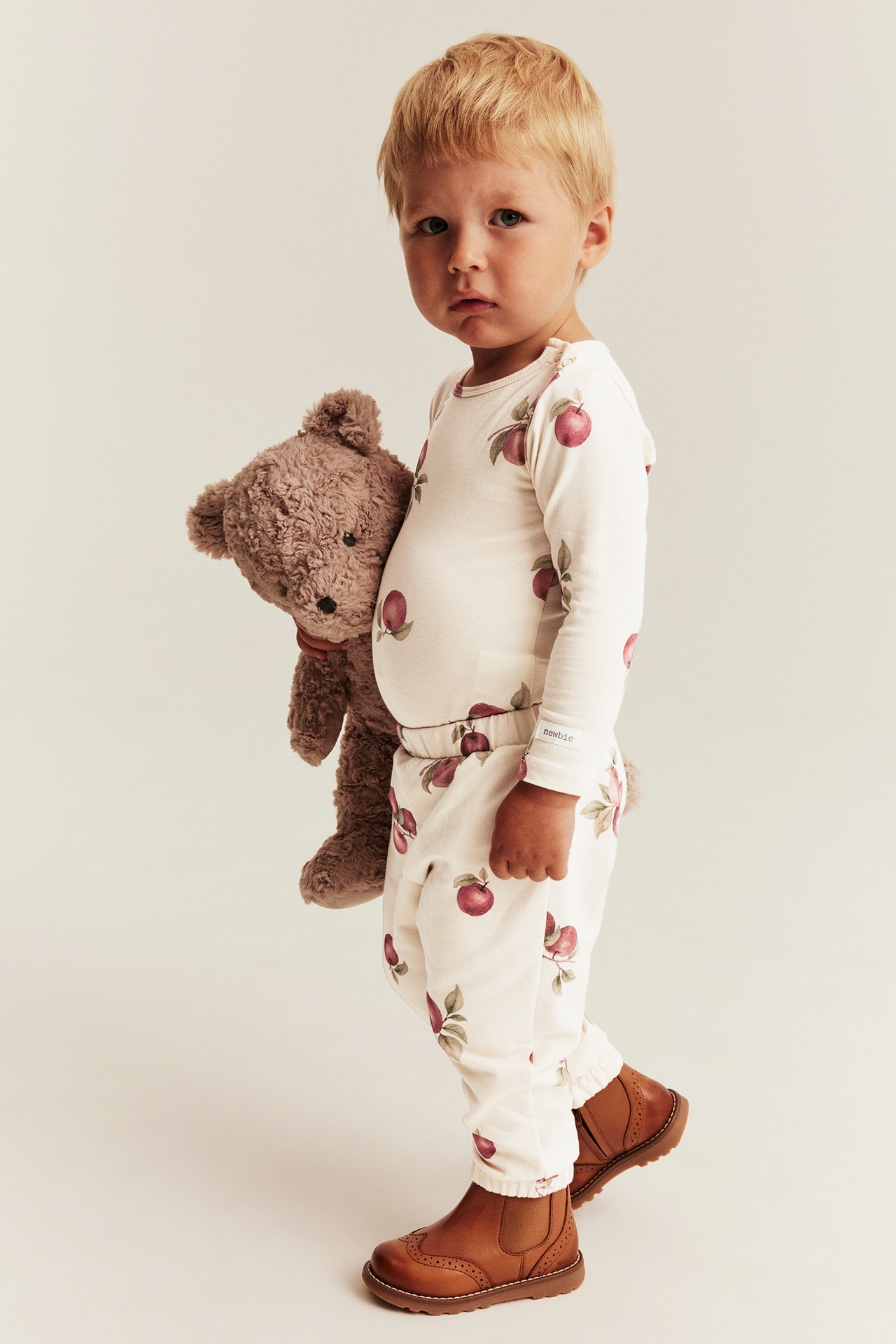 Barns off-white eple-sett, topp og bukser, brun bamse, brune Chelsea-boots, sideprofil.