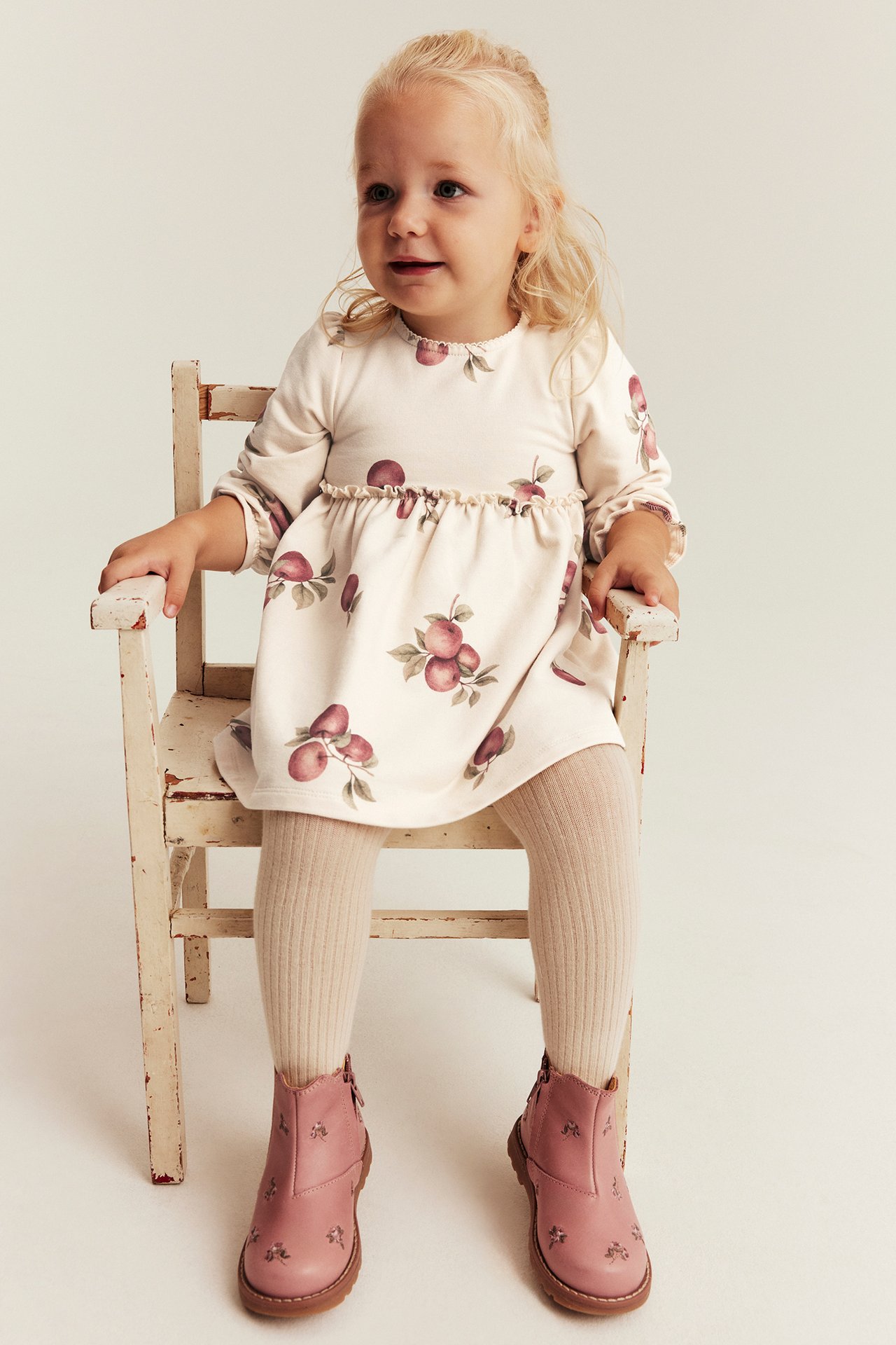 Framsida av en babyflicka i beige klänning med äppeltryck, ribbade beiga strumpbyxor och rosa broderade ankelstövlar.