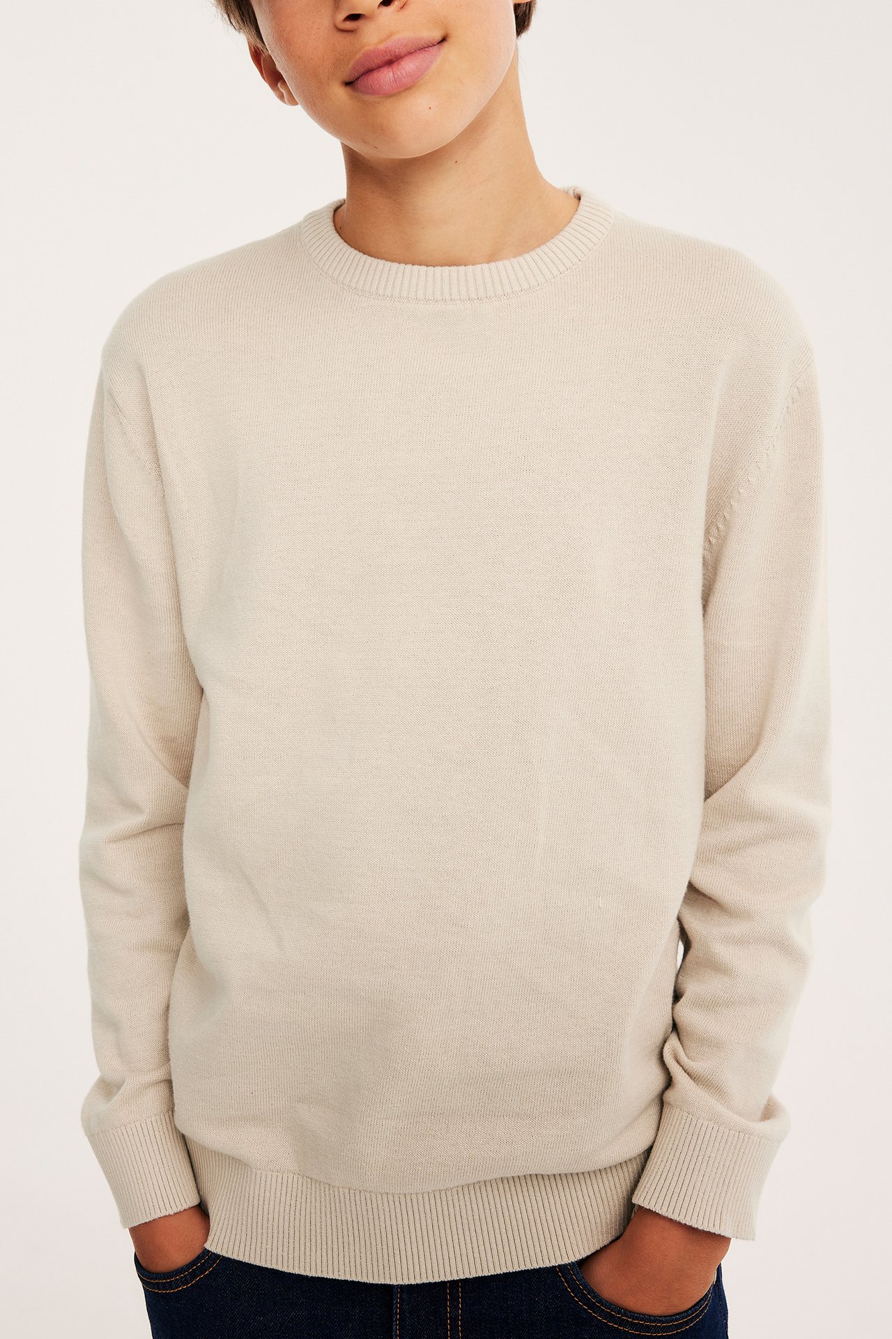 Forfra av et barn i beige strikket genser med rund hals og mørkeblå jeans.
