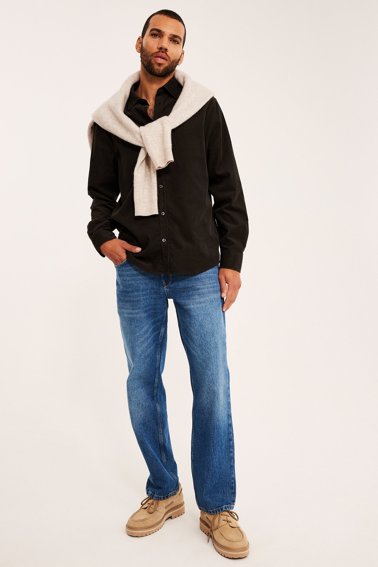 Framsida av en man i svart långärmad skjorta, beige stickad tröja, blå jeans och bruna mockaskor.