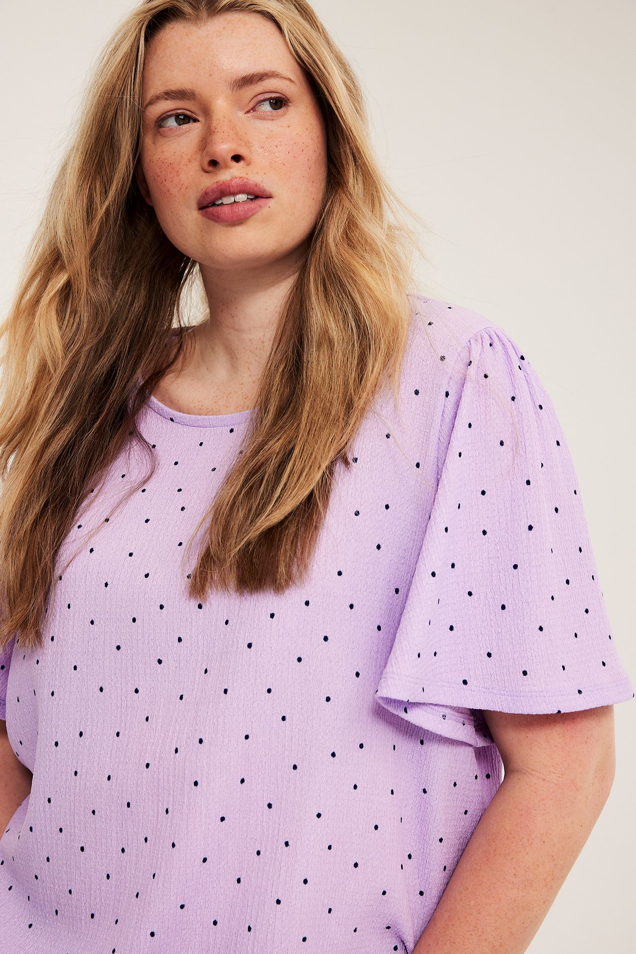 Närbild framifrån av en kvinna i en ljuslila prickig kortärmad topp med volangärmar.