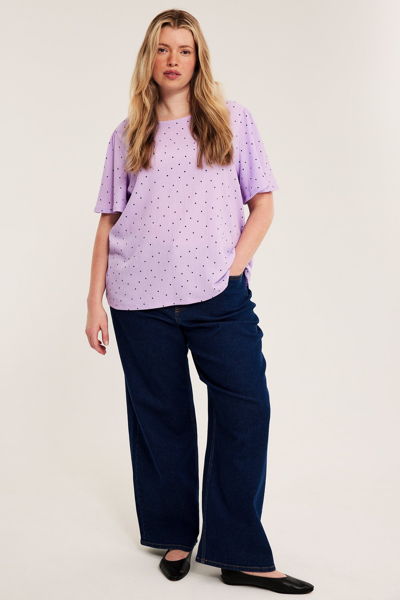 Frontvy av kvinna i ljuslila prickig kortärmad topp, mörkblå vida jeans och svarta ballerinaskor.