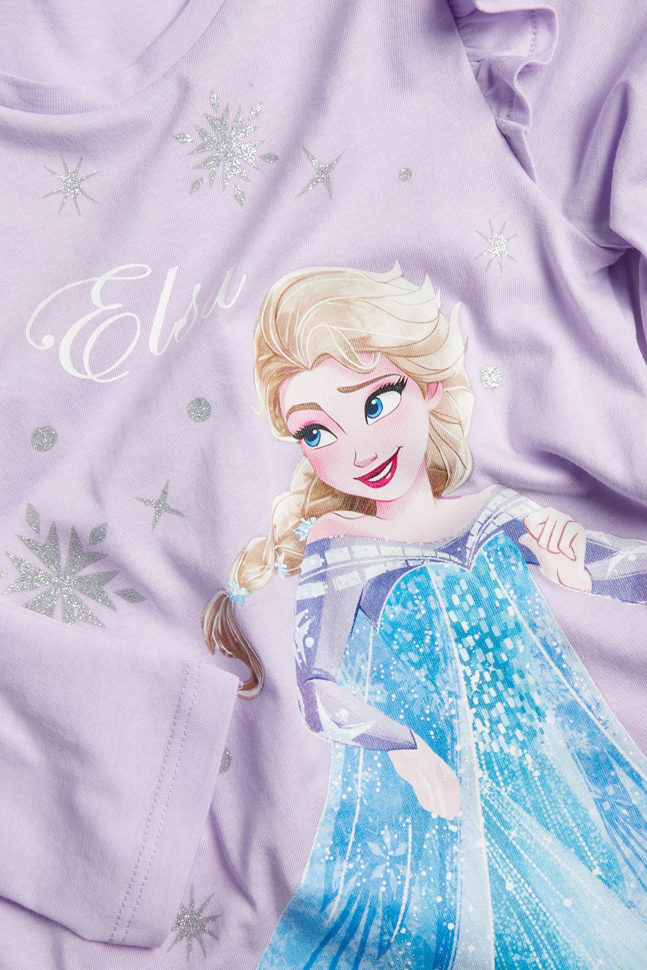Lähikuva lasten laventelinliilasta pitkähihaisesta paidasta, jossa Elsa-painatus ja glitteriä.