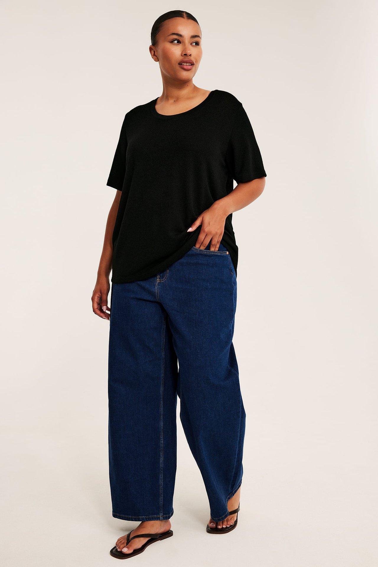 Framifrån: kvinnlig modell i en svart ledig t-shirt, blå vida jeans och svarta flip-flops.