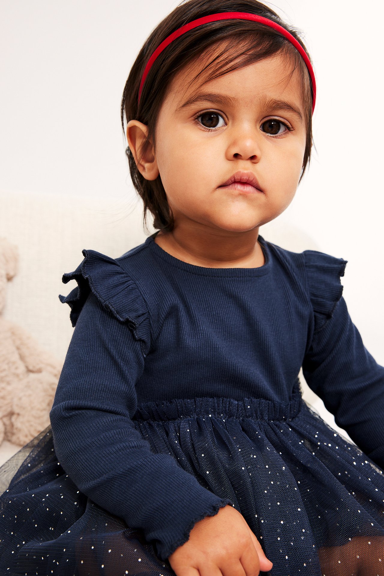 Framsida: Baby i mörkblå ribbad långärmad klänning med prickig tyllkjol och rött hårband.