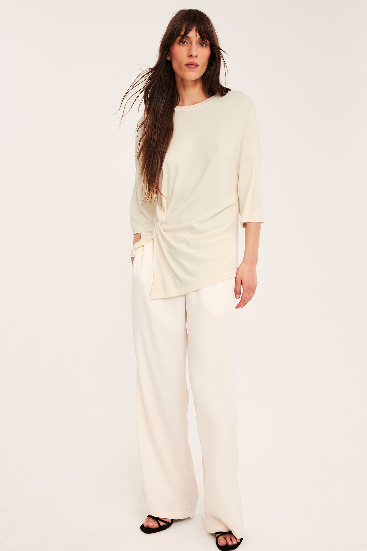 Framsida: kvinnlig modell i beige topp med knutdetalj, matchande byxor och svarta sandaler.