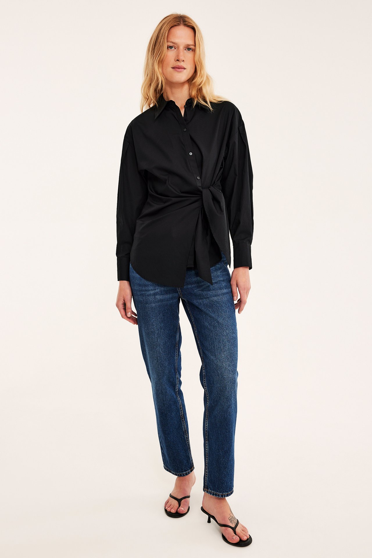 Frontbilde av en kvinne i svart knyteskjorte, blå jeans og svarte lavhælte tåsandaler.