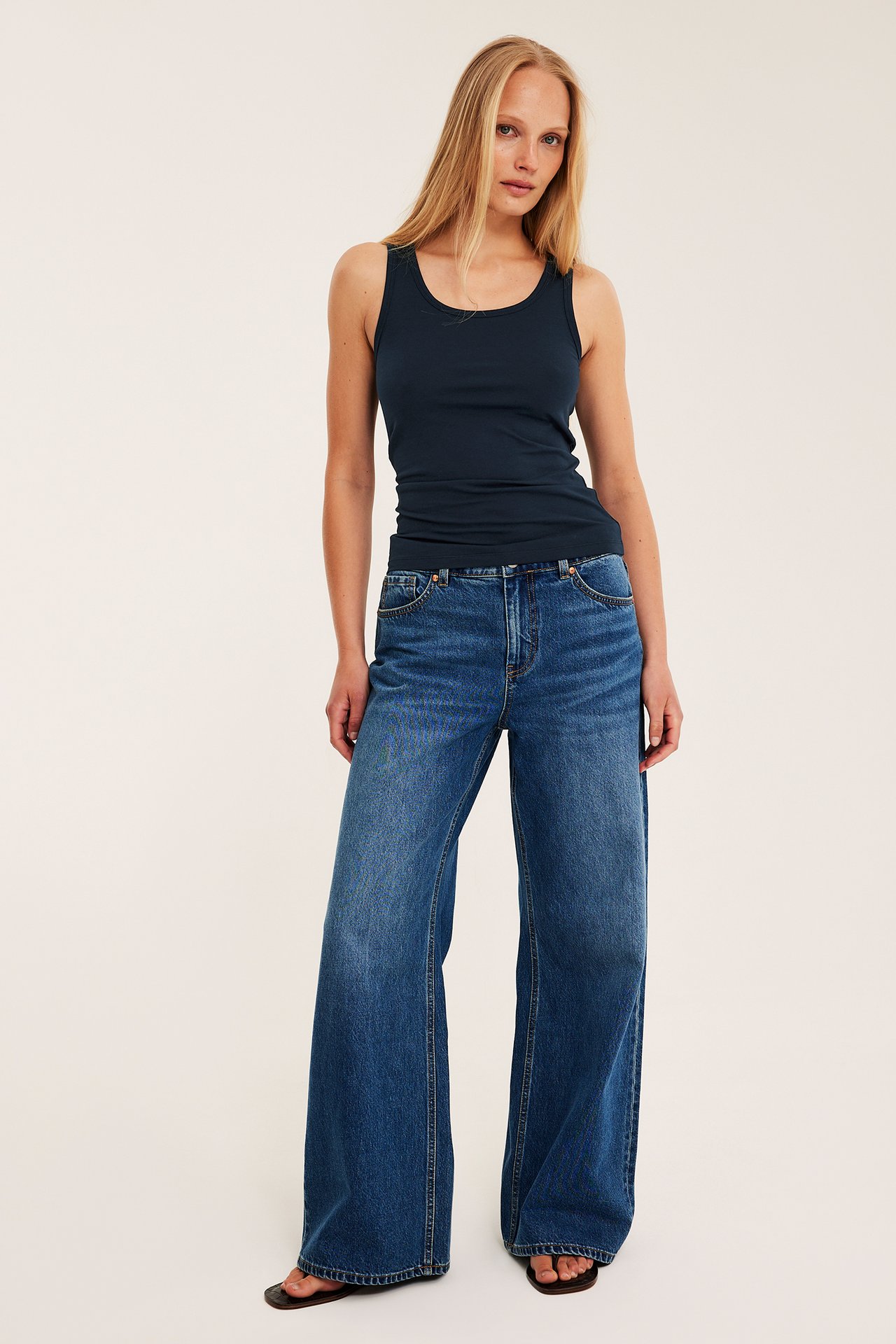 Edestäpäin: nainen tummansinisessä puuvillatopissa, sinisissä leveissä farkuissa ja tummissa sandaaleissa.
