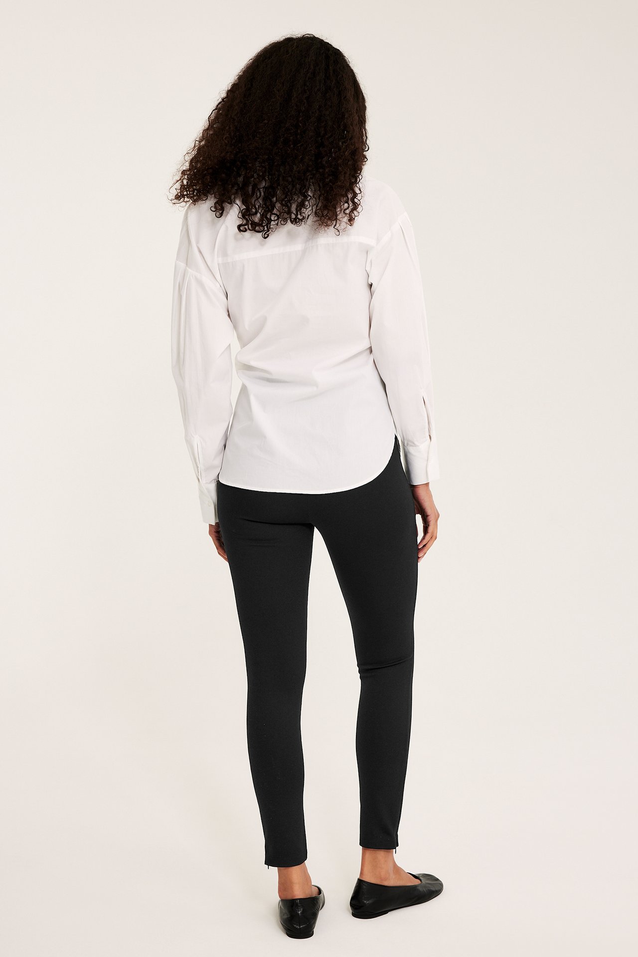 Bakfra av en kvinne i svarte leggings med glidelås ved anklene, en hvit skjorte og svarte ballerinasko.