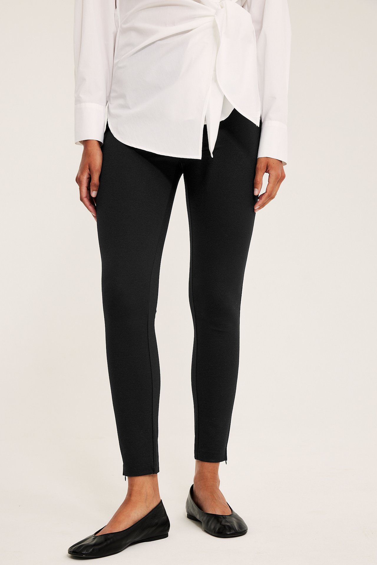 Frontbilde av kvinne i svarte, smale leggings med glidelåser ved anklene, en hvit omslagsskjorte og svarte flate sko.