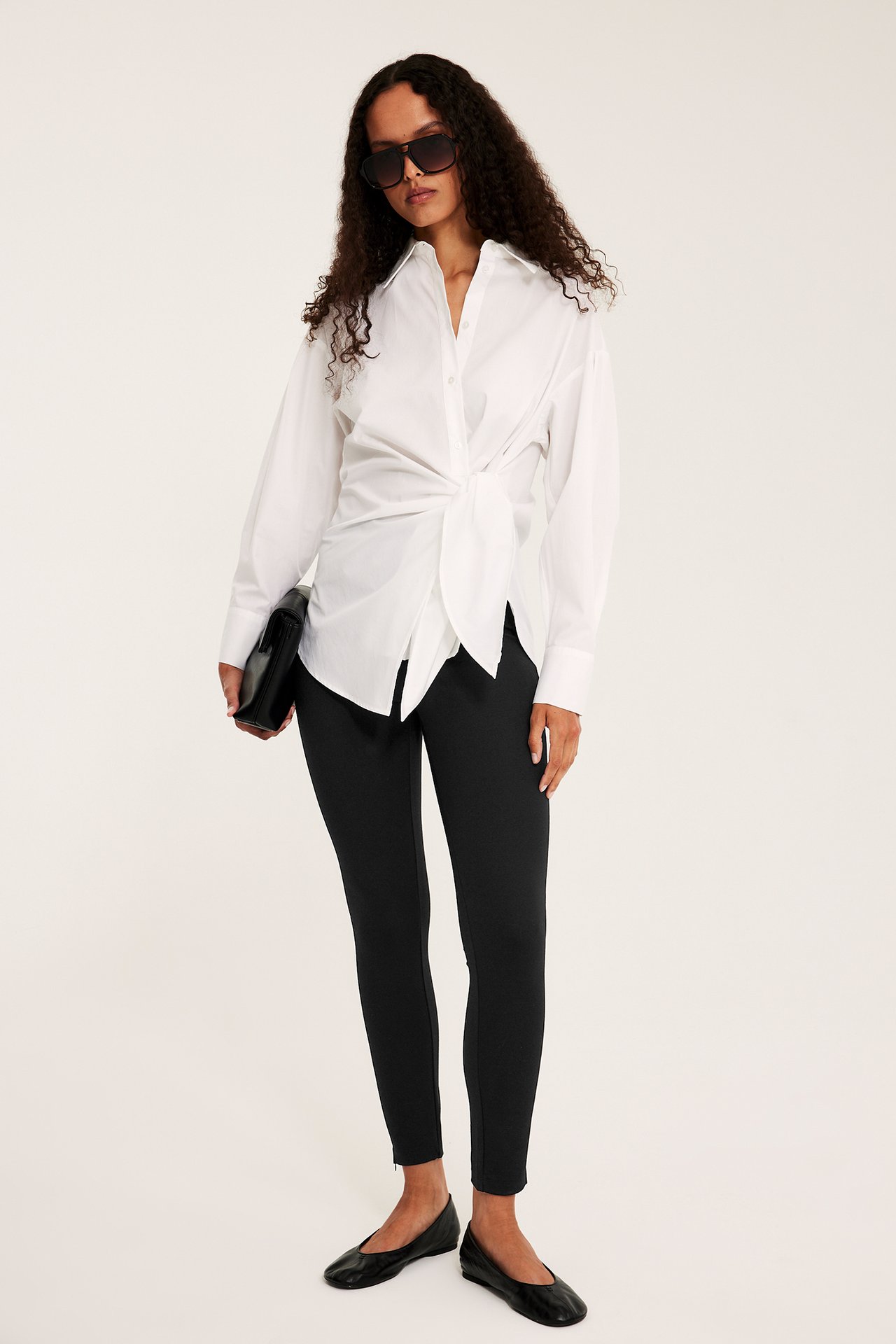 Frontvisning av kvinne i svarte leggings med glidelåser ved anklene, hvit knyttet skjorta, svarte ballerinasko, holder clutch.