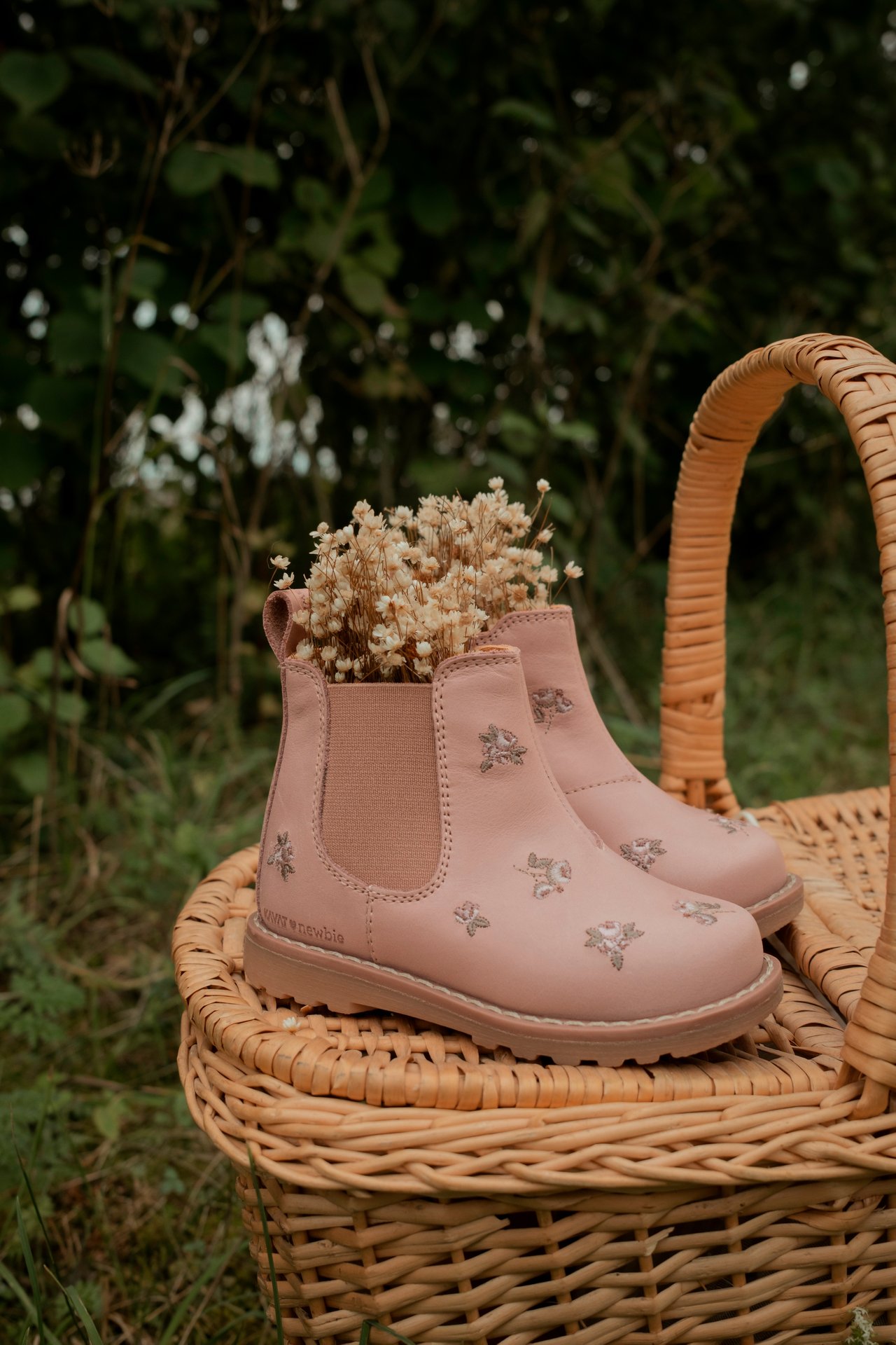 Nærbilde av rosa barne-chelseaboots med blomsterbroderi, tørkede blomster, på kurv.