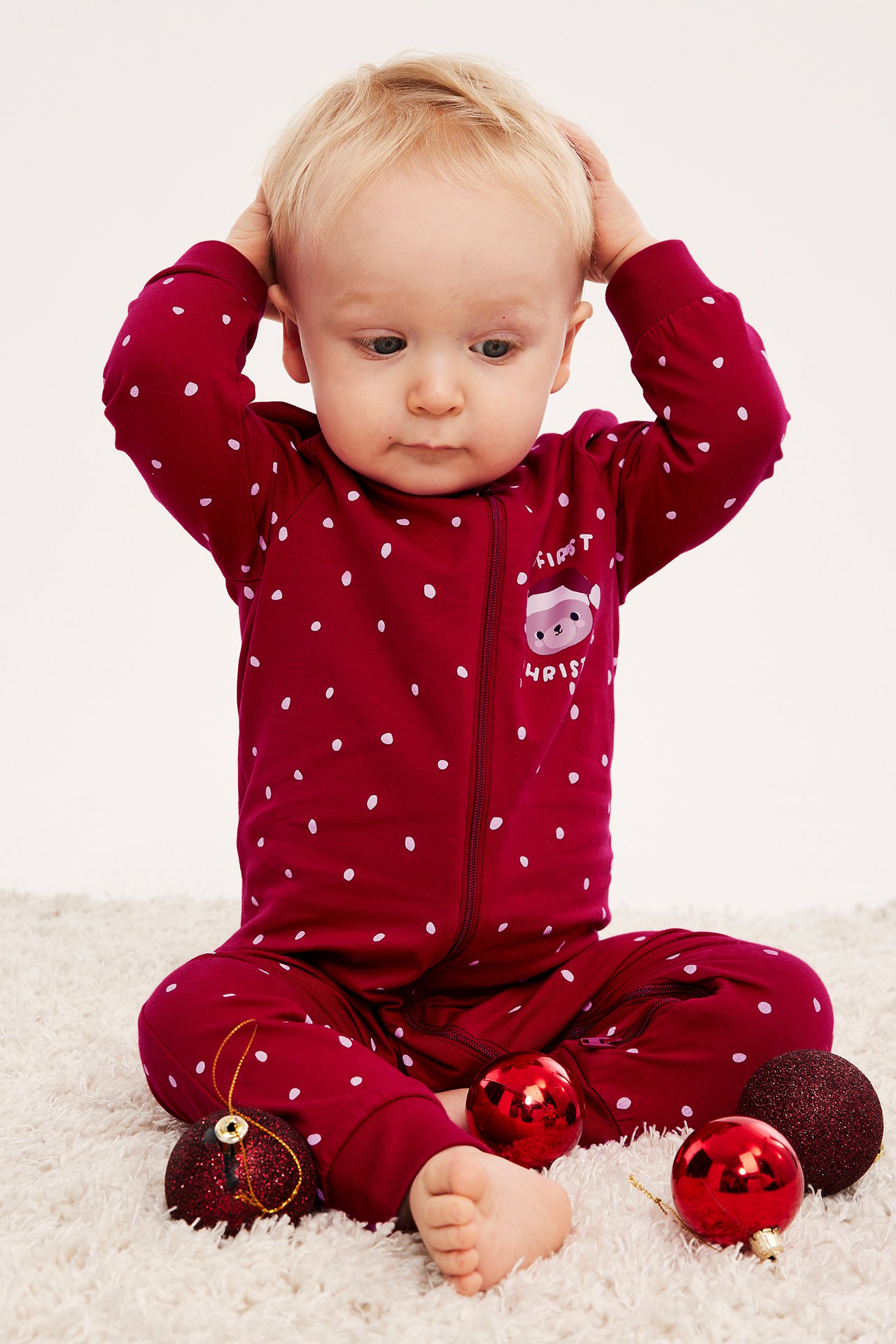 Front vy av en baby i röd prickig pyjamas med "First Christmas"-tryck och dragkedja.