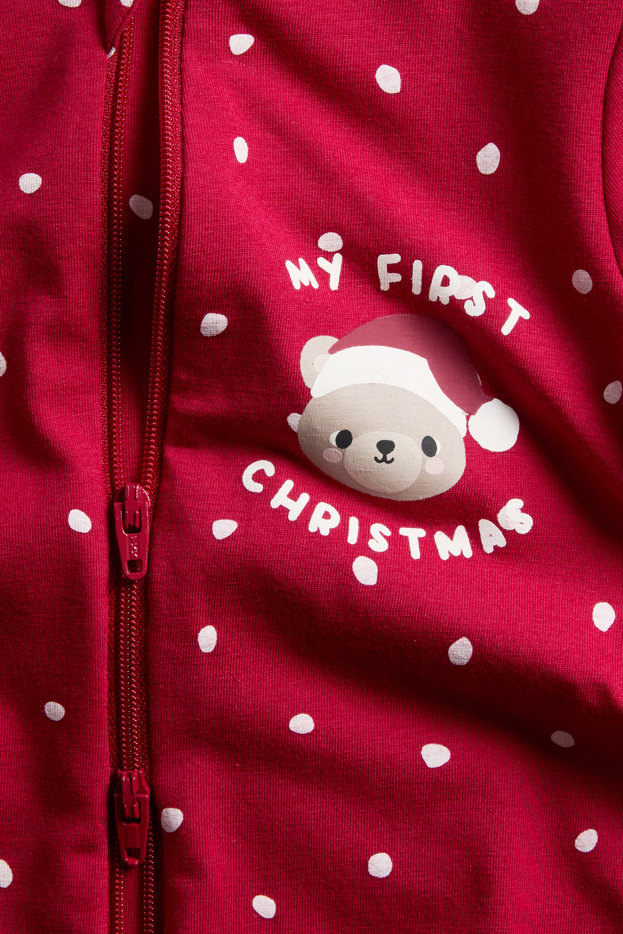 Närbild på röd babydräkt med vita prickar, ett 'My First Christmas' björnmotiv och dragkedja.