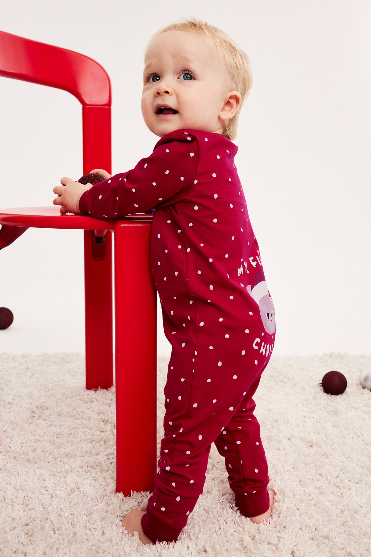 Sidovy av en baby i en vinröd prickig julpyjamas, hållande en mörk prydnad.
