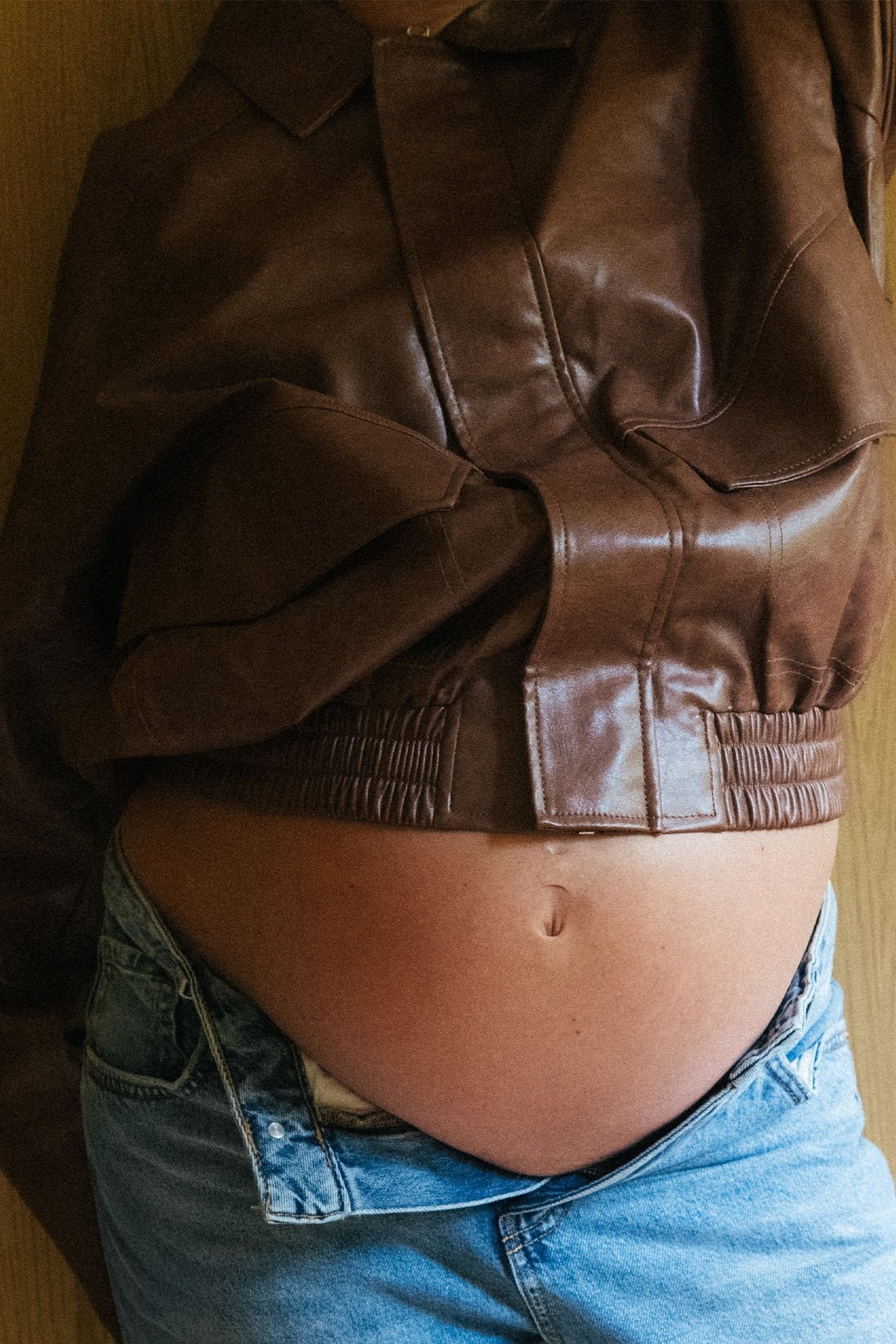 Framifrån, gravid kvinna i brun läderbomberjacka över ouppknäppta ljusblå jeans.