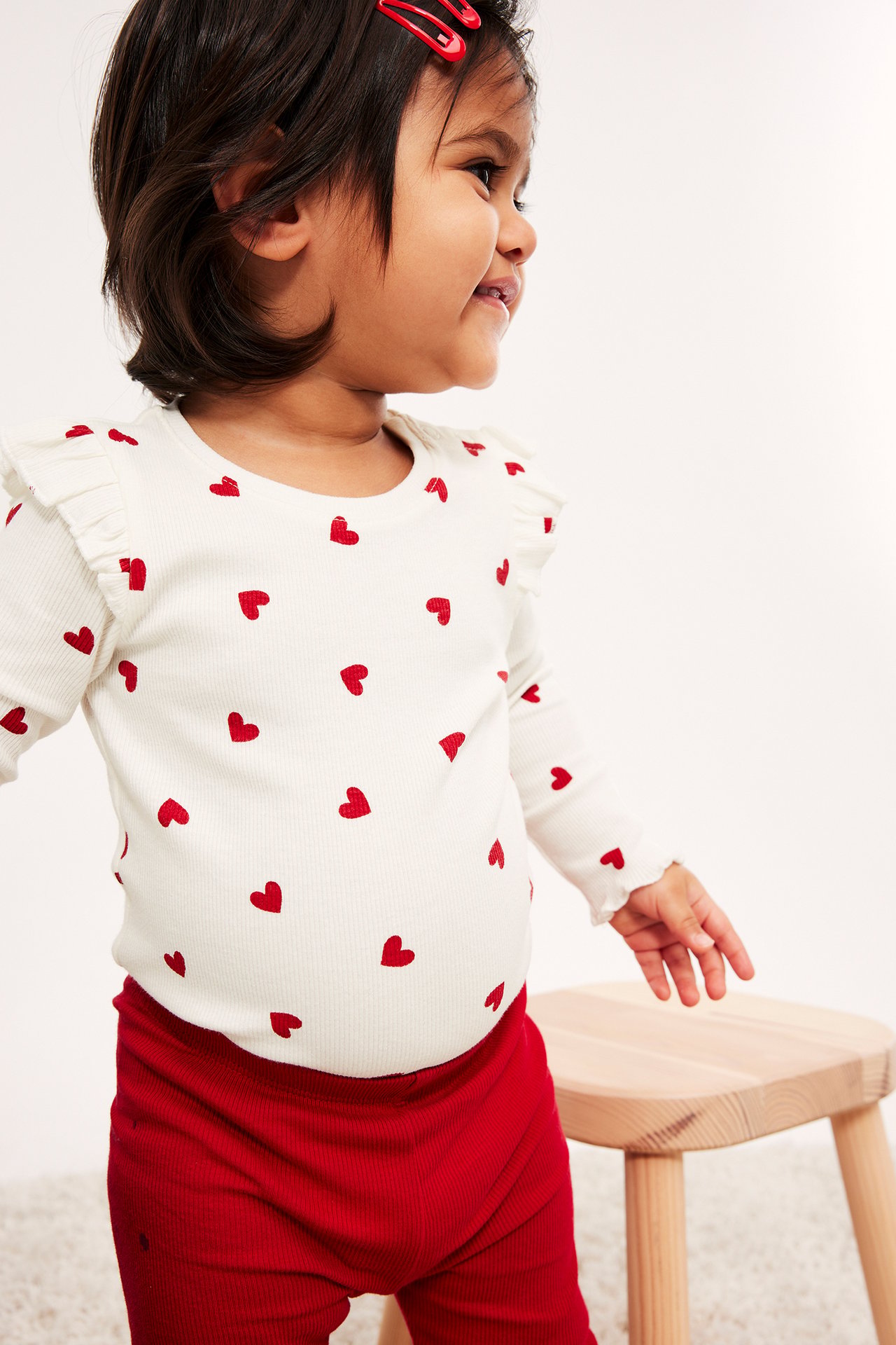 Sidovy av baby i vit långärmad ribbad topp med röda hjärtan och röda ribbade leggings.