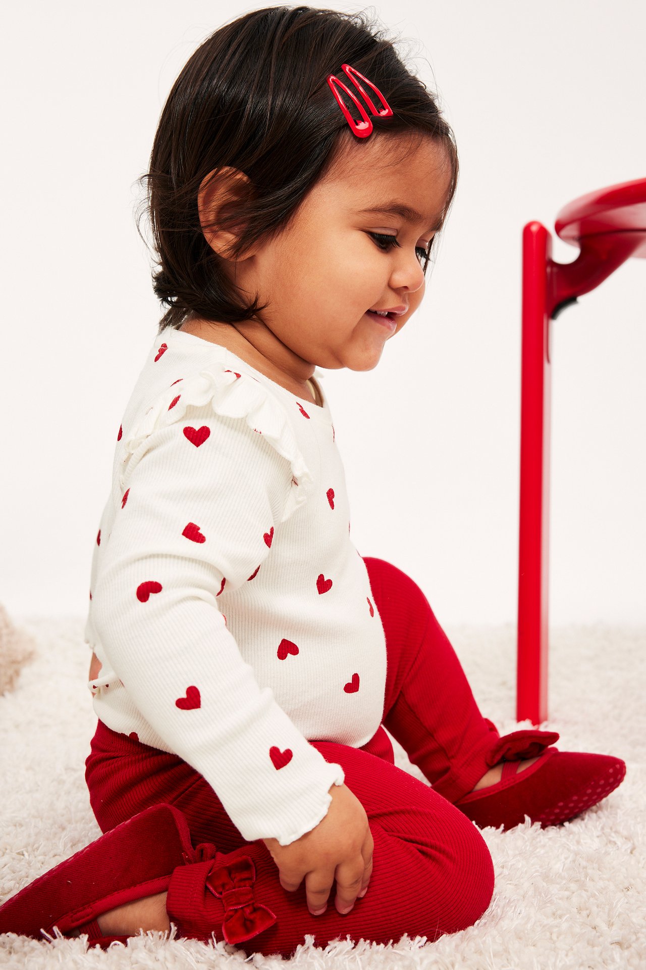 Sidovy: bebis bär vit hjärtmönstrad topp, röda ribbade leggings, röda sammetsskor med rosett, röda hårspännen.