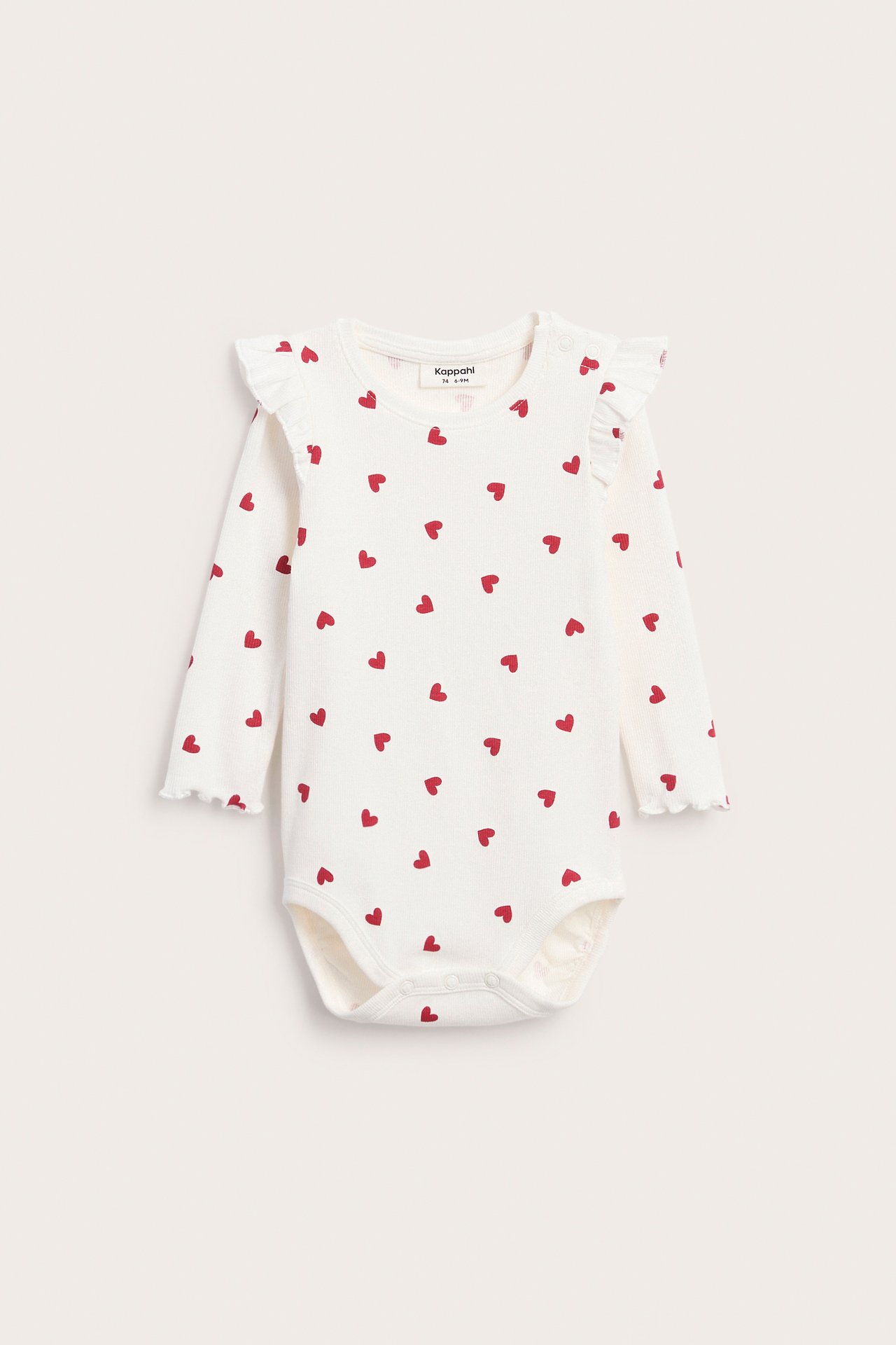 Framifrån: Vit långärmad babybody med röda hjärtan och volanger på axlarna.