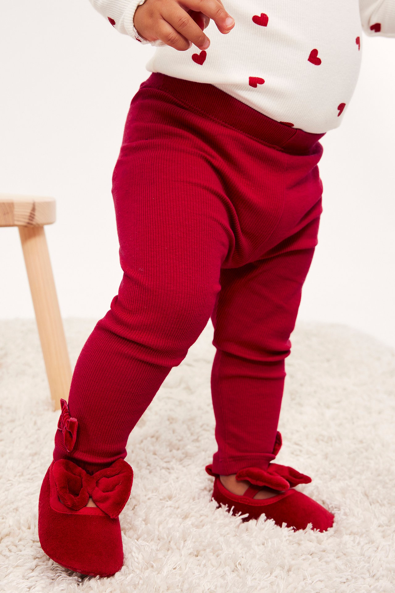 Bebis bär röda ribbade leggings, röda sammetsskor med rosetter, och en vit topp med röda hjärtan.