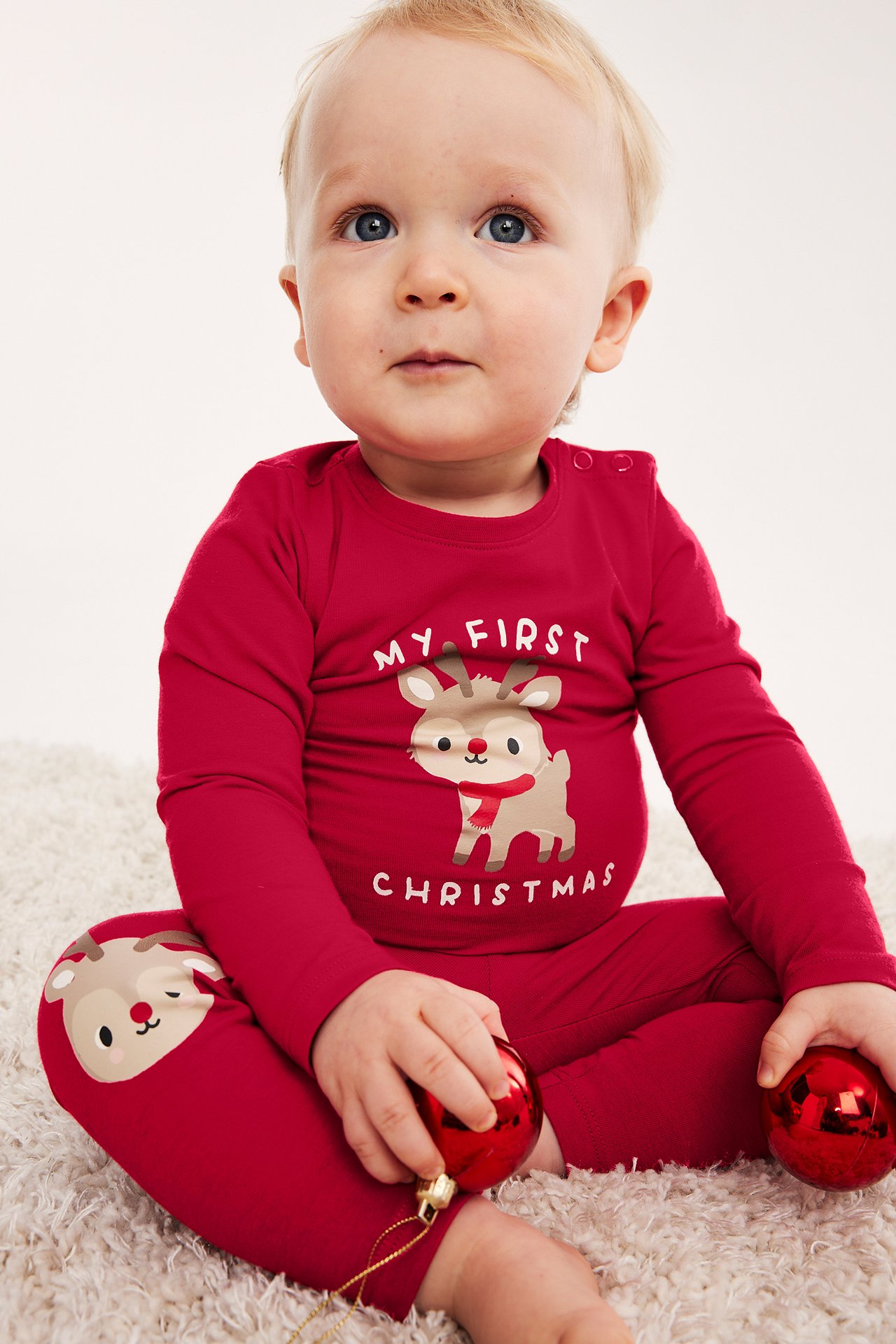 Dziecko w czerwonej bluzce i spodniach 'My First Christmas' z reniferem, trzymające czerwone bombki.