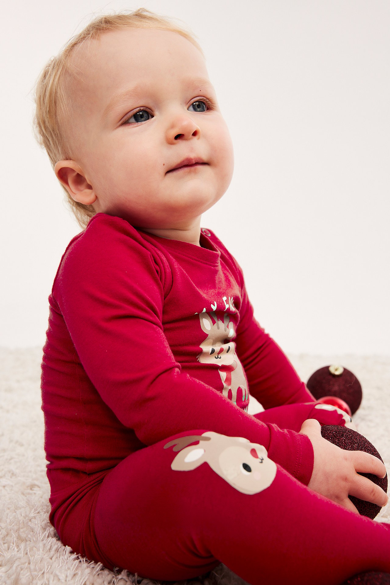 Baby i röd pyjamas med renprint håller en glittrig röd julkula.