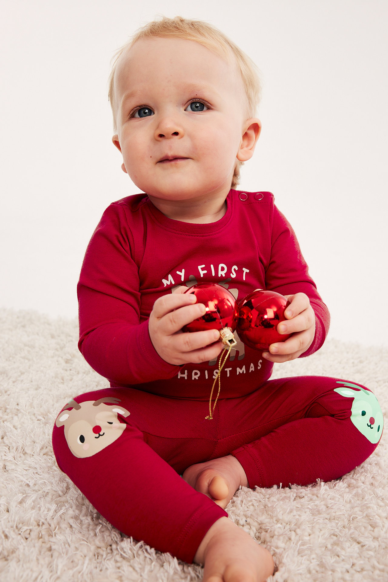 Framifrån: Baby i röd 'My First Christmas'-pyjamas med renknän, håller två röda julkulor.