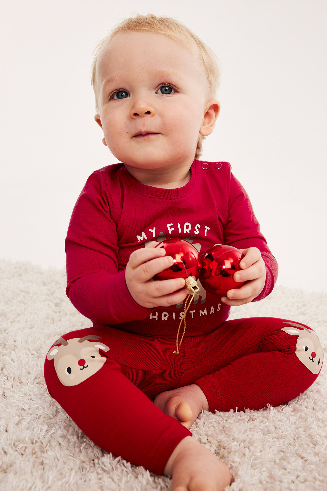 Baby i rød 'My First Christmas'-genser og reinsdyr knepatch leggings, holder to røde julekuler.