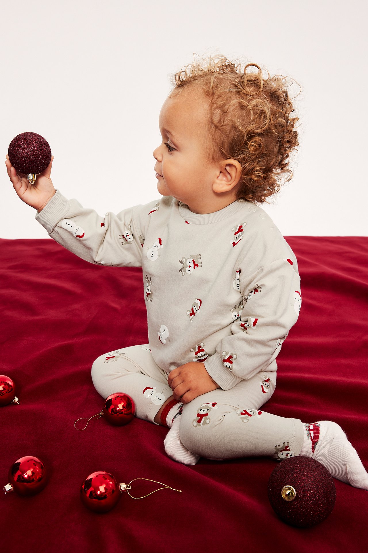 Sidovy av ett baby som bär ett beiget pyjamasset med jultryck och håller en röd glitterkula.
