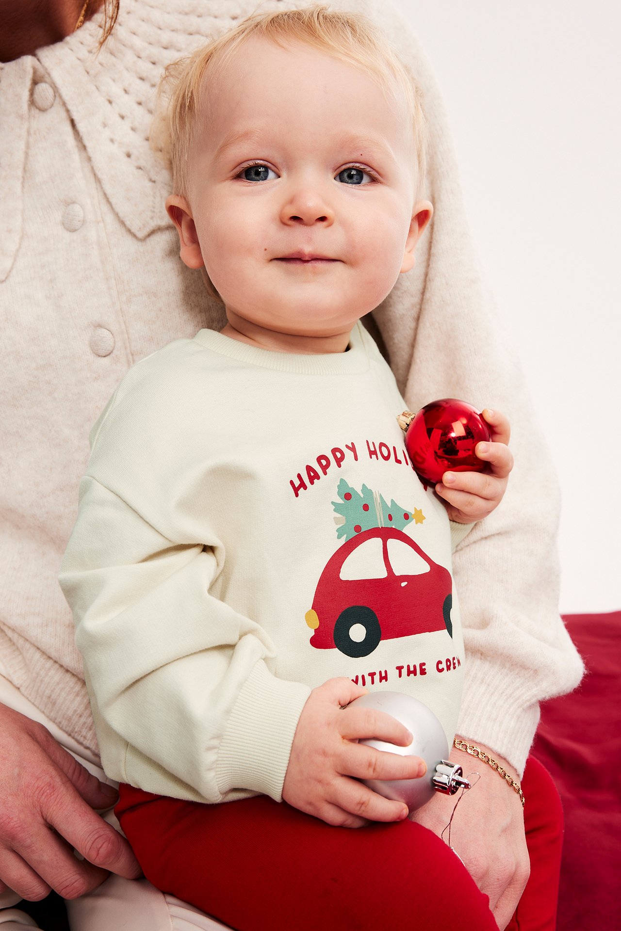 Framifrån, baby i en krämvit tröja med julmotiv och röda byxor, håller i julgranskulor.