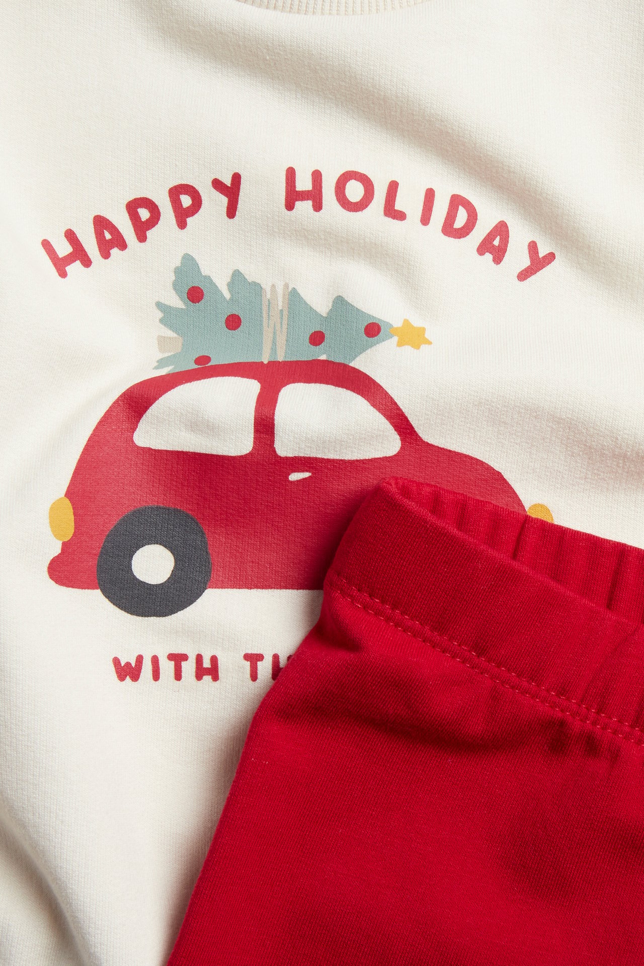 Närbild av en barns krämvita tröja med "Happy Holiday"-tryck med röd bil och röda leggings.