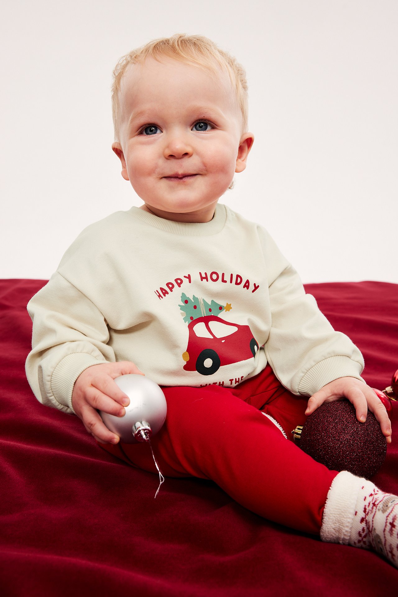 Baby i en beige 'Happy Holiday'-tröja, röda byxor och vita strumpor, håller i julgranskulor.