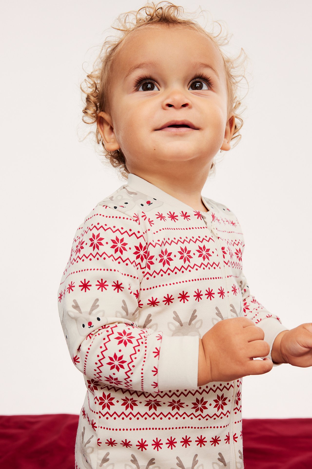 Framifrån, baby i en ljusbeige pyjamas med dragkedja, rött snöflinge- och grått renmönster.