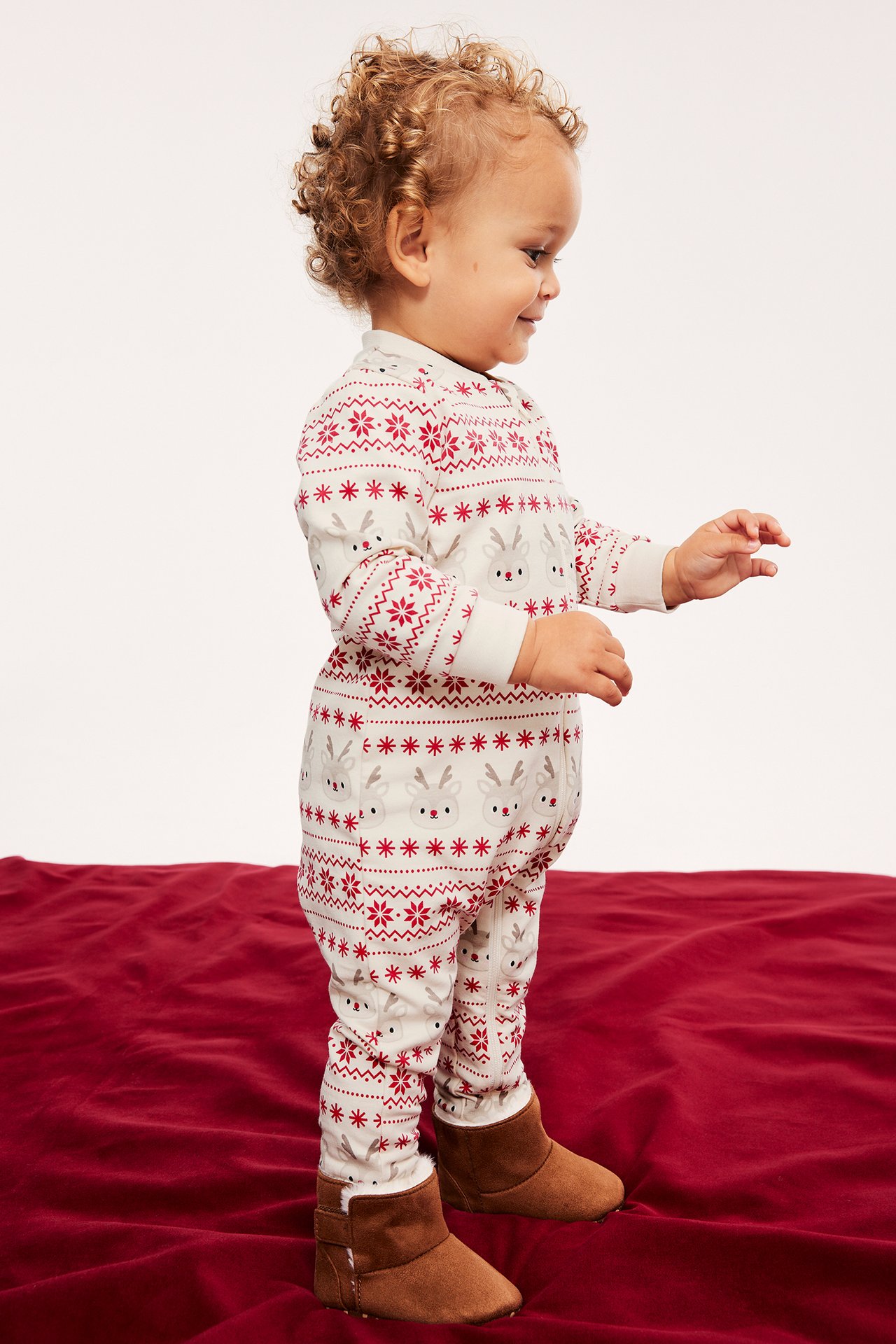 Sidovy av baby i benvit pyjamas med renmönster och bruna stövlar på en röd filt.