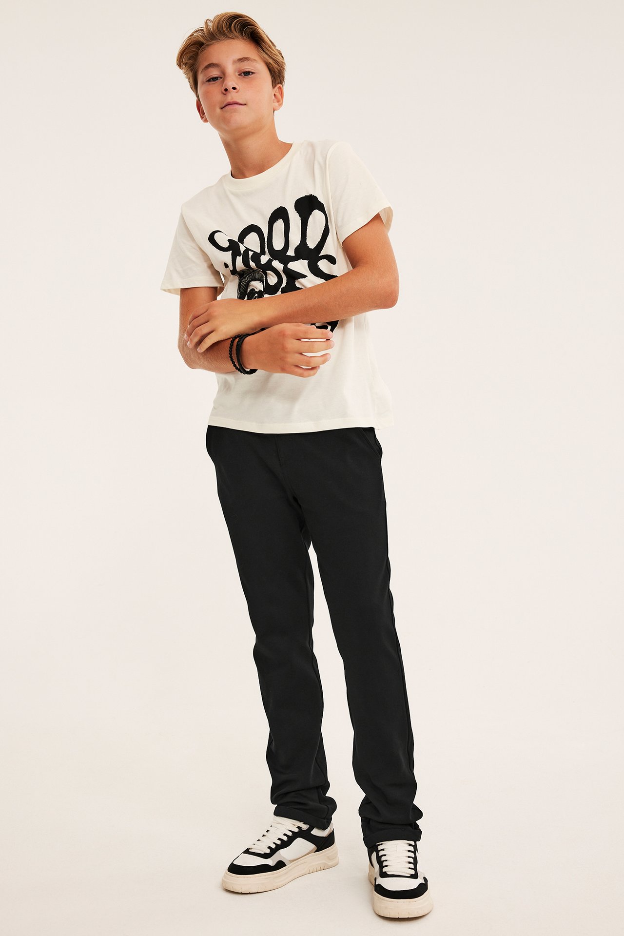 Forfra, barn i en kremfarget 'Good Vibes' t-skjorte, svarte bukser, hvite sneakers, svarte armbånd.