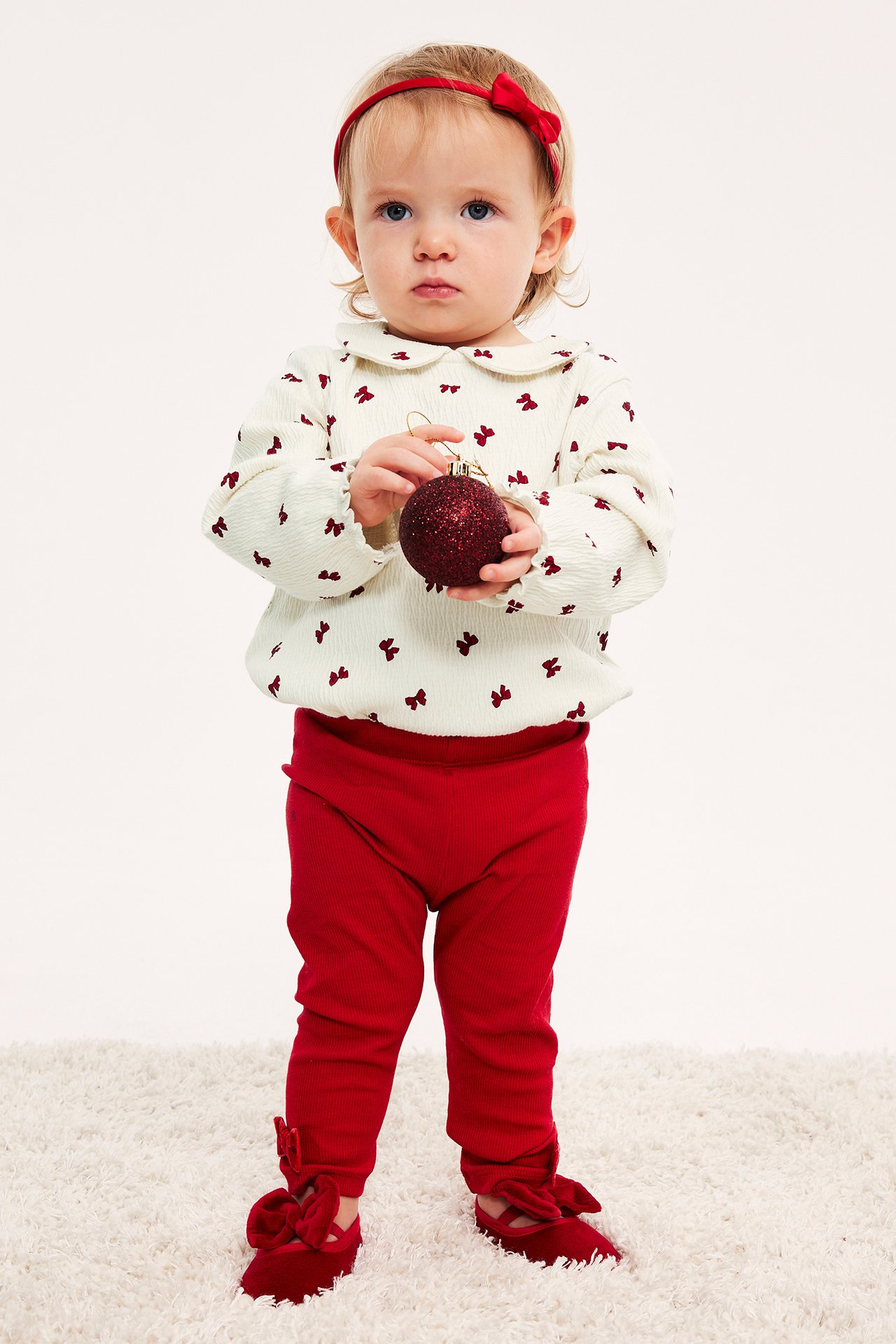 Baby i cremevit topp med röda rosetter, röda leggings, röda rosettskor, rött diadem.