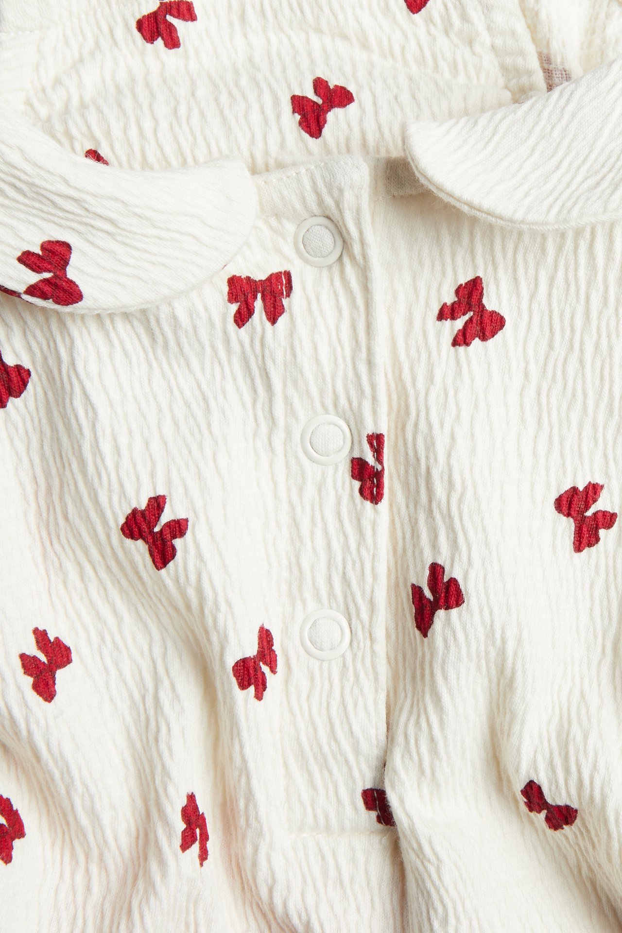Närbild på baby overall i krämig textur med röda rosetter, rund krage och tryckknappar.