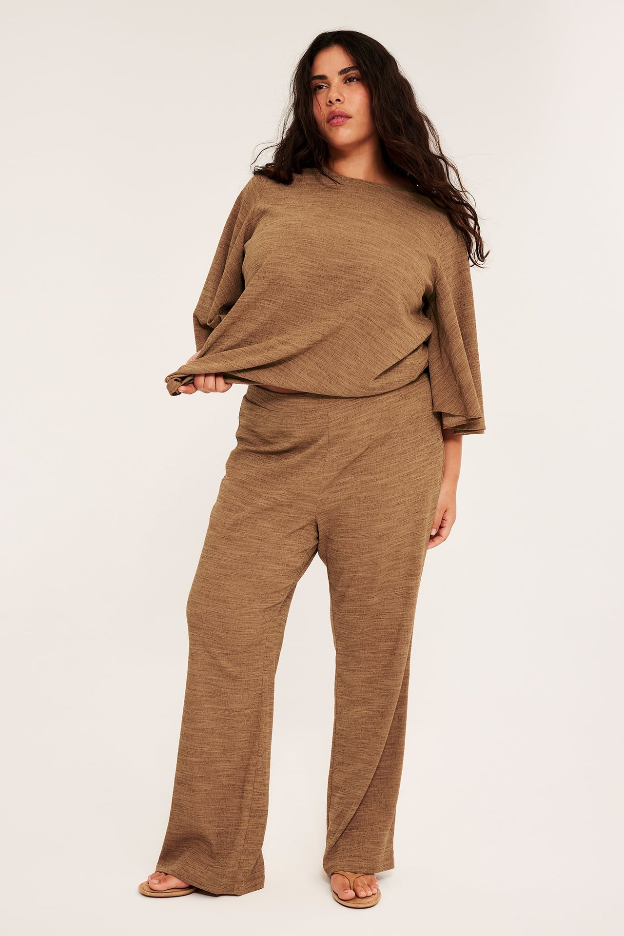 Nainen edestä ruskeissa bootcut-trikoohousuissa, löysässä topissa ja vaaleanruskeissa sandaaleissa.