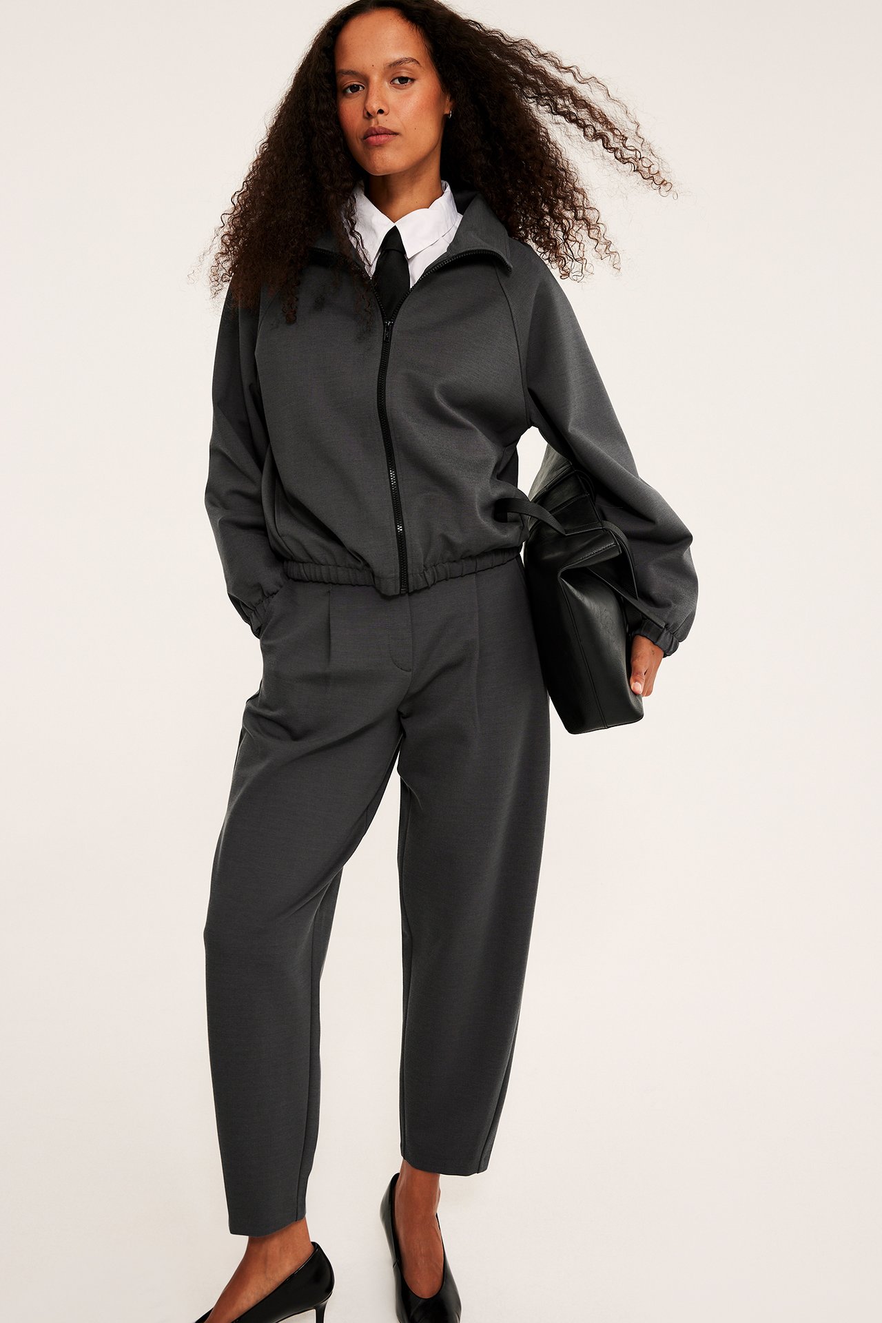 Front vy: Mörkgrå zipjacka och matchande cropped byxor, vit skjorta, svart slips, pumps, hållen väska.
